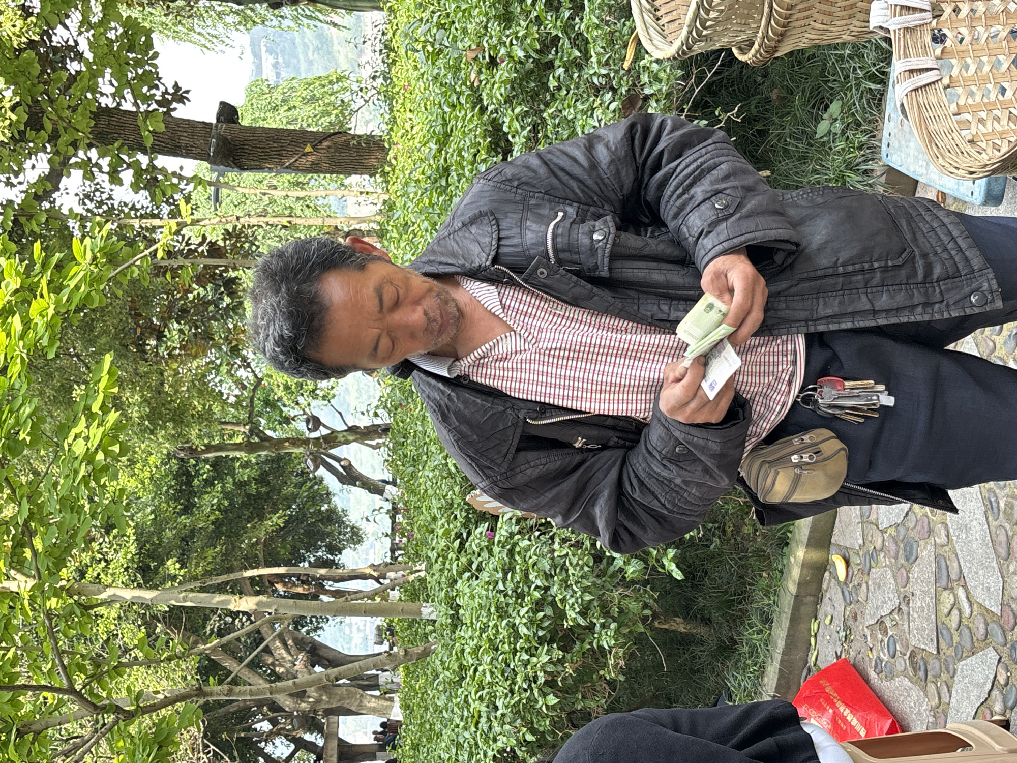 An Asian man in a black jacket and red and white checkered shirt stands outside near trees and bushes counting money in his hands. A pouch with keys hangs from his waist. Wicker baskets sit to his right. The location is Jiangjin, China.
