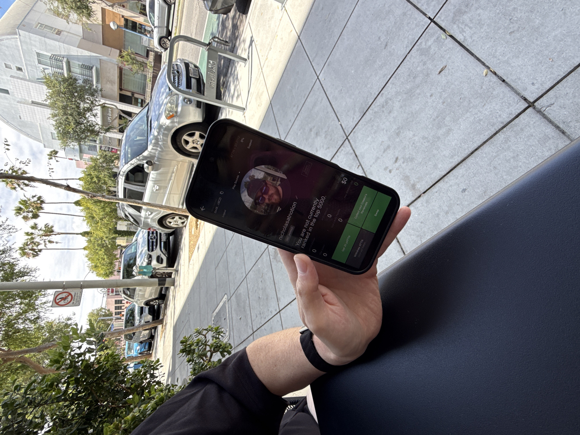 Here is a forensic description of the image:
A person holds a smartphone displaying an app profile identified as "nicholasatnotion." The screen indicates the user is "not currently ranked in the top 5000" and displays a balance of $0. The person is wearing a black shirt and a black fitness tracker on their wrist, and the phone is resting on a black surface.
The background reveals an outdoor scene with palm trees and a building with a "UPS Store" sign. There are several cars parked along the street, and a "No Bicycles" sign is visible. The setting appears to be a city street in the United States of America. Based on the architectural style, palm trees, and signage observed, the location is likely West Hollywood.