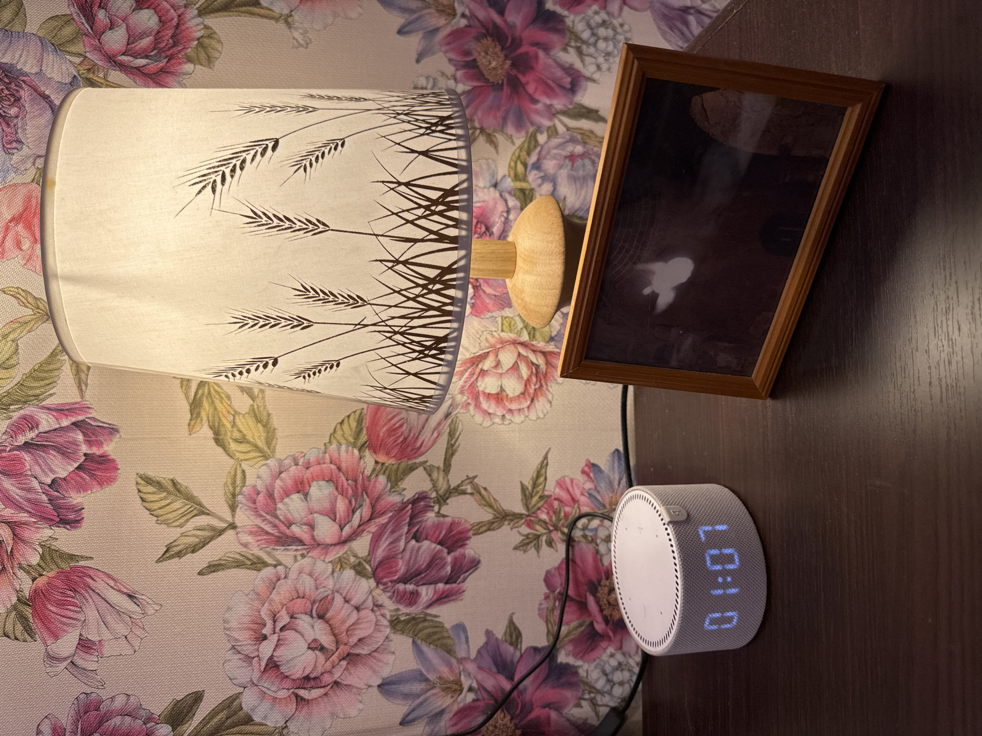The image depicts a close-up, eye-level shot of a bedside table. The table surface is dark brown and appears to be made of wood. On the table, to the left, is a white, cylindrical smart speaker with a textured surface and visible speaker holes. A blue digital clock on the front of the speaker displays "01:07". To the right of the speaker, slightly behind it, is a framed photograph. The frame is made of wood with a light brown finish and is positioned at an angle, showing a dark image within. Behind the objects, the wall is covered in floral wallpaper featuring large, pink and purple flowers and green leaves. A lamp with a white lampshade, decorated with a pattern of wheat stalks, is visible in the background, its wooden base resting on the table. A black electrical cord runs from the smart speaker towards the left edge of the frame. There are no people or animals present in the scene. The lighting suggests an indoor setting, possibly a bedroom at night. The image does not provide any specific visual cues to indicate the location of Almaty, Kazakhstan.