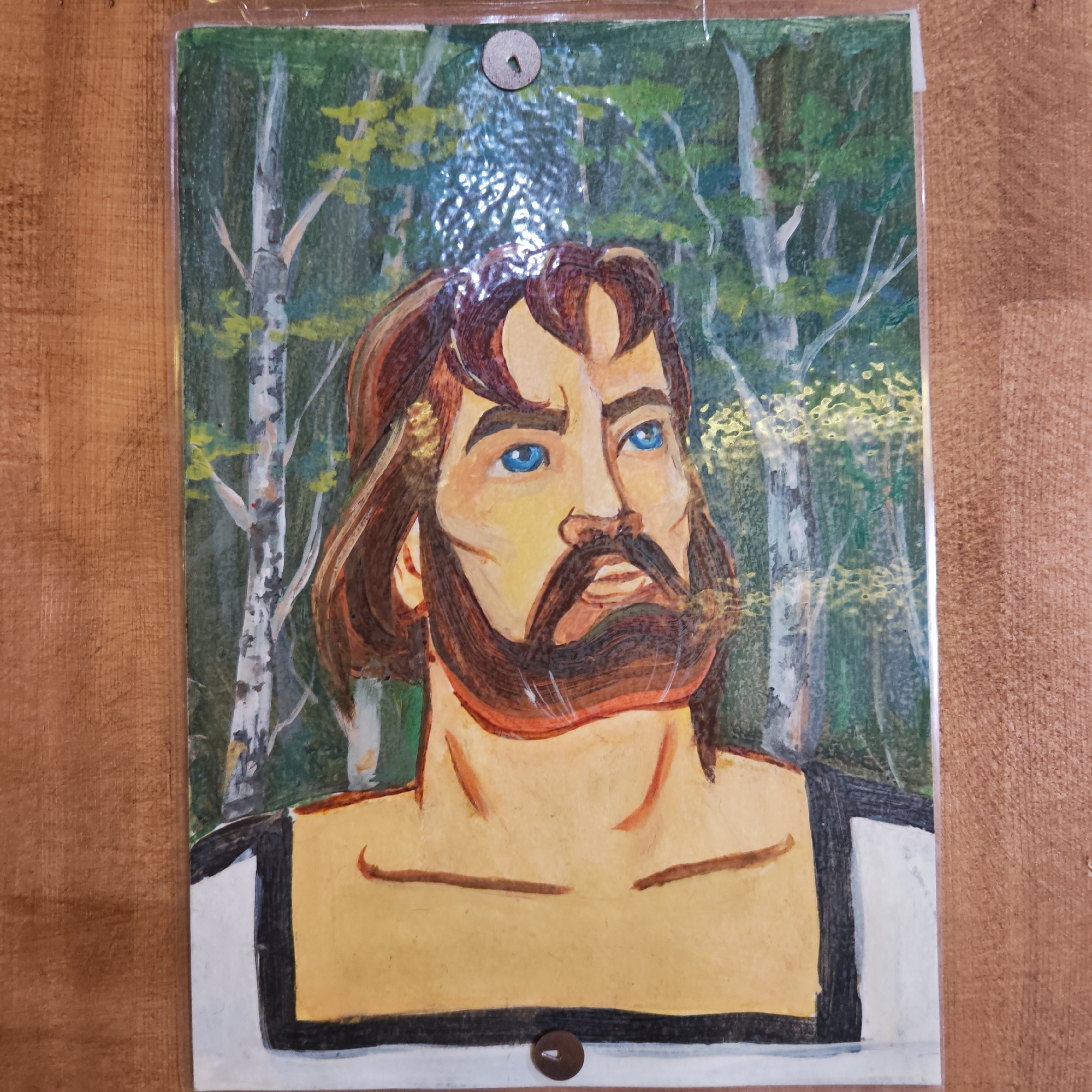 A framed portrait painting hangs on a wooden wall. The painting depicts a man with brown hair, a beard, and striking blue eyes, looking upwards and to the right. He is dressed in a white shirt with black trim. The background is a green forest with birch trees. The artwork is encased in a plastic sleeve, which is attached to the wall with two metal tacks, one at the top and one at the bottom of the frame. There is no information available to confirm that this image was taken in Novo-Nikol'skoe, Russia.