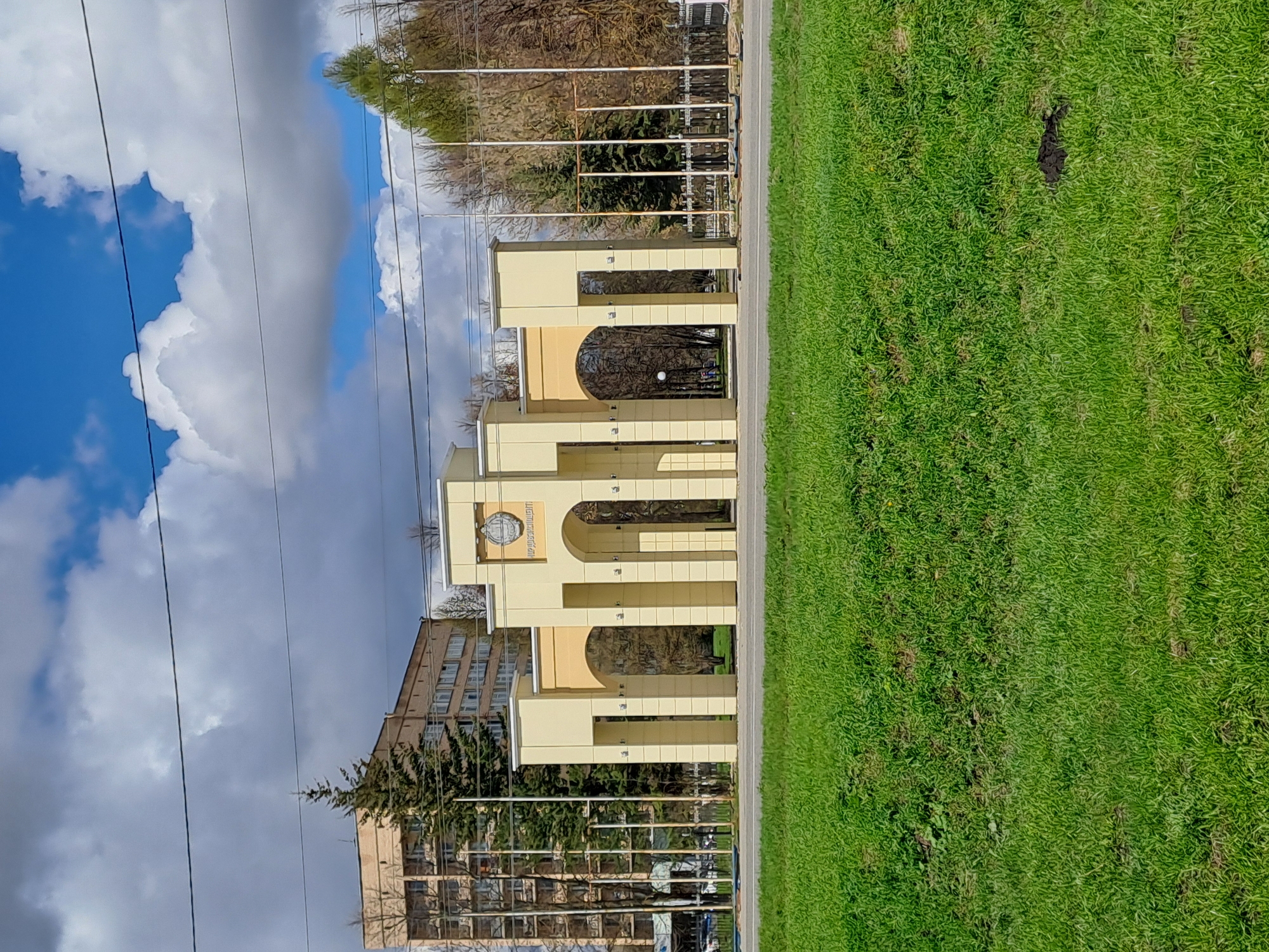 This horizontally oriented image depicts an outdoor scene featuring an entrance to a university campus in Yalga, Russia, under a partly cloudy sky.
The central subject is a prominent light yellow or beige archway structure with multiple arched passages, likely serving as a gateway or ceremonial entrance. Near the top of the central arch, a circular emblem or seal is visible. The text on this emblem, written in Cyrillic, clearly reads "МОРДОВСКИЙ ГОСУДАРСТВЕННЫЙ УНИВЕРСИТЕТ" (Mordovian State University), confirming the location.
To the right of the archway, a wide expanse of vibrant green grass fills the foreground, showing a few darker patches of soil. A paved road or path separates the grass from the archway and the other architectural elements. Behind the archway, part of a multi-story building with numerous windows, presumably a university building, can be seen. Further in the background, a mix of bare deciduous trees and some evergreen trees are visible. Several tall, slender flagpoles are also present behind the archway.
The sky, occupying the left side of the frame, presents a dynamic mix of clear blue patches and large, white, fluffy cumulus clouds, along with some grayer clouds. Several dark utility lines or wires stretch across the sky. The bright illumination suggests it is daytime, likely late morning or early afternoon. No people or specific activities are observed in the static scene. The image provides a clear view of the university entrance and its immediate surroundings.