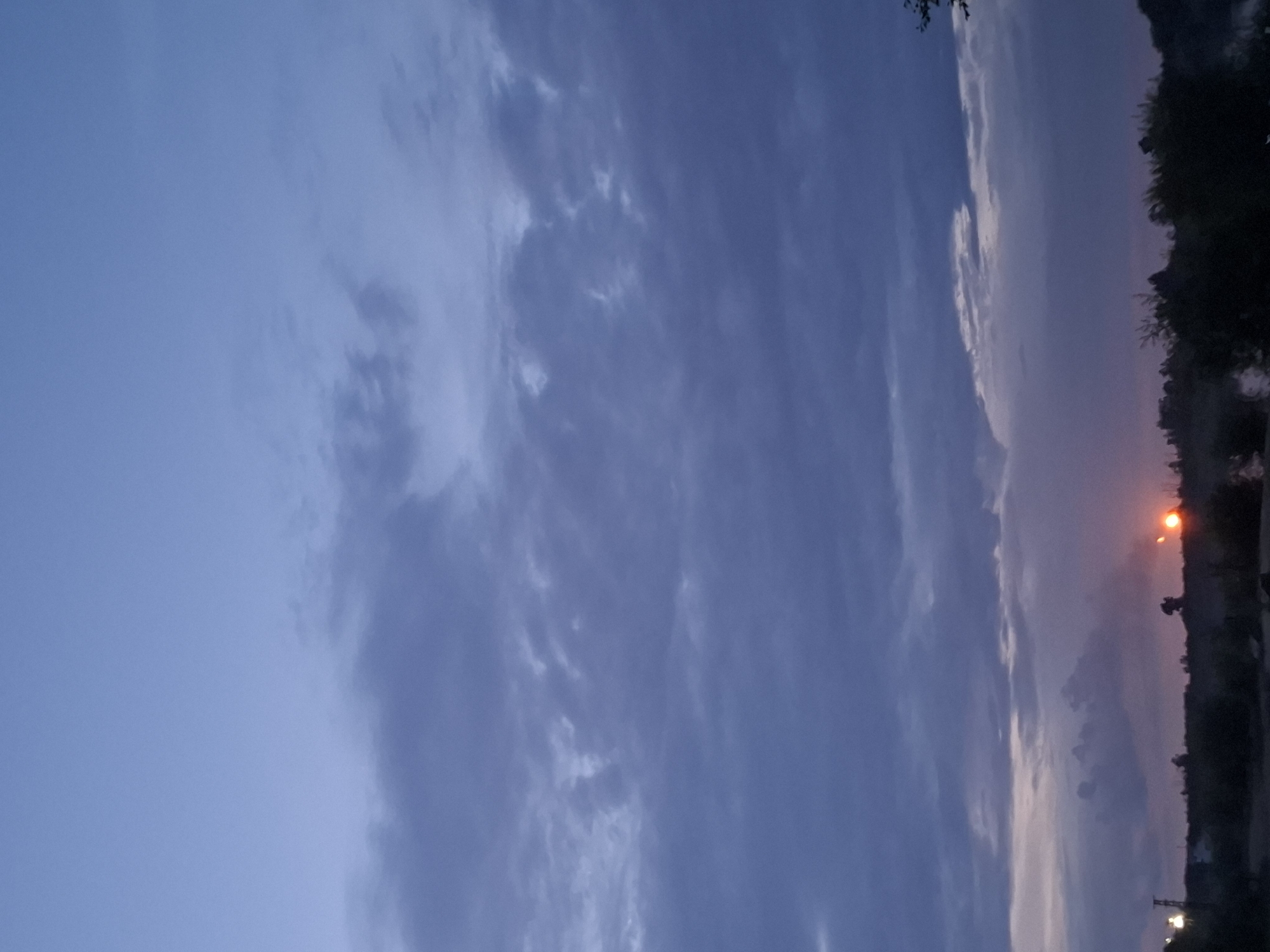The image depicts a twilight scene with a vast, overcast sky transitioning from a deep blue at the top to lighter hues near the horizon. Dark, textured clouds dominate the upper and middle portions of the frame. Along the horizon, a silhouetted landscape of trees and low-lying vegetation is visible. A bright, orange orb, likely the setting sun or artificial light, is visible near the right side of the horizon, casting a faint glow. In the distance, on the left side, a tall, thin structure resembling a utility pole with lights is present. The overall atmosphere is serene and subdued, characteristic of dusk. The location is specified as Svobodny, Russia.