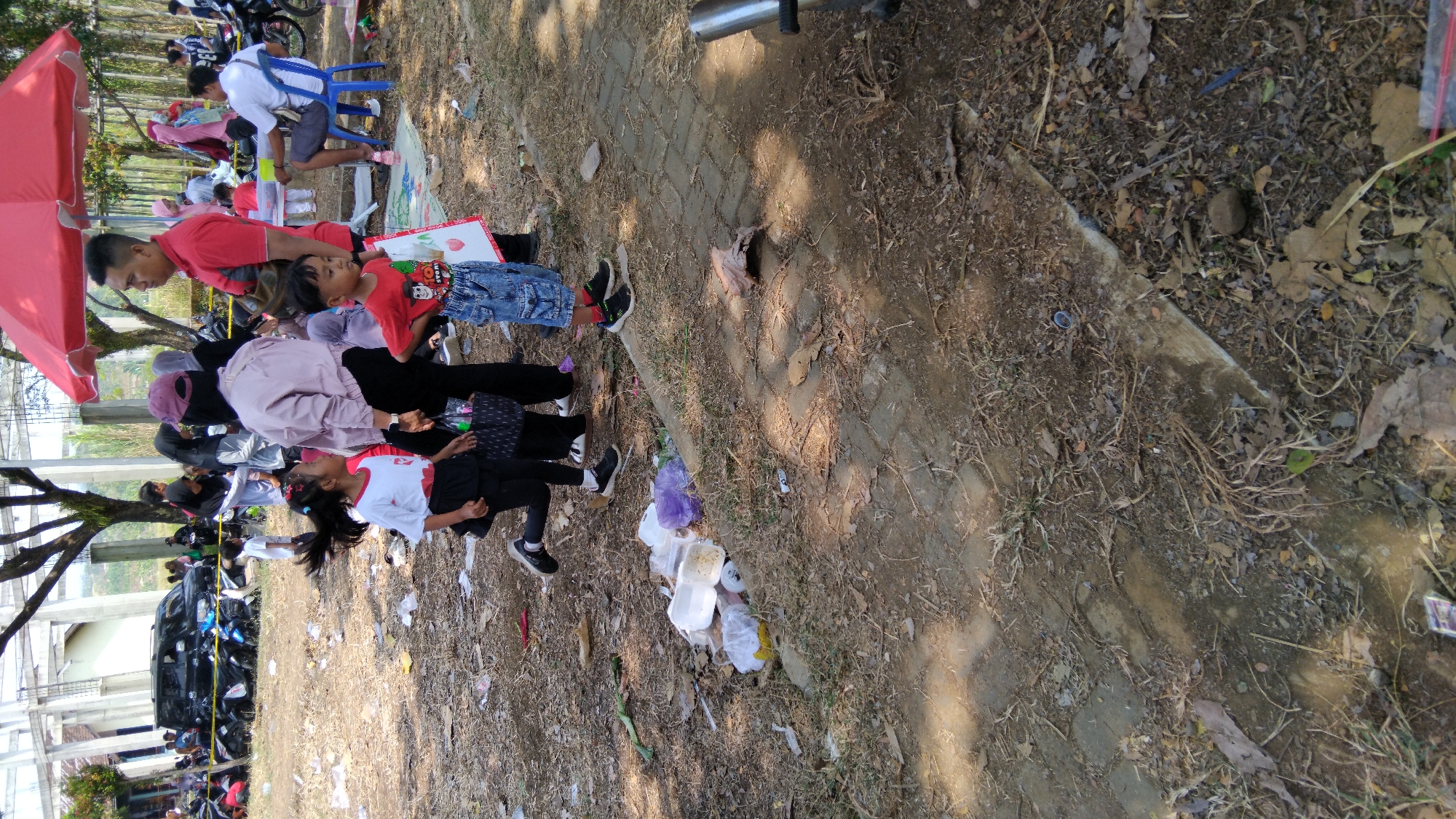 The image depicts a sunny outdoor scene with a gathering of people. In the foreground, two young children are the primary focus. A girl, wearing a white t-shirt with red accents and a black skirt, is kneeling and looking to the left. Beside her, a boy in a red t-shirt with a cartoon graphic and denim shorts stands facing forward, holding a white paper.

Behind them, a group of adults and children are visible. One adult man in a red shirt stands to the right of the children, and further back, a woman in a light purple jacket and a burgundy headscarf is seen. Several other individuals, some in headscarves, are scattered throughout the background. A red umbrella is partially visible at the top right, suggesting a vendor or recreational setup.

The environment appears to be a park or open public space with a dirt and paved ground. There is a significant amount of litter scattered on the ground, including plastic containers, bags, and leaves, indicating a lack of cleanliness. In the background, a black SUV is parked among several motorcycles, and a light-colored building structure with visible pillars is present. The presence of a red umbrella and casual attire suggests a public event or market setting. The overall scene appears to be within the city of Posong, Indonesia, given the context provided.