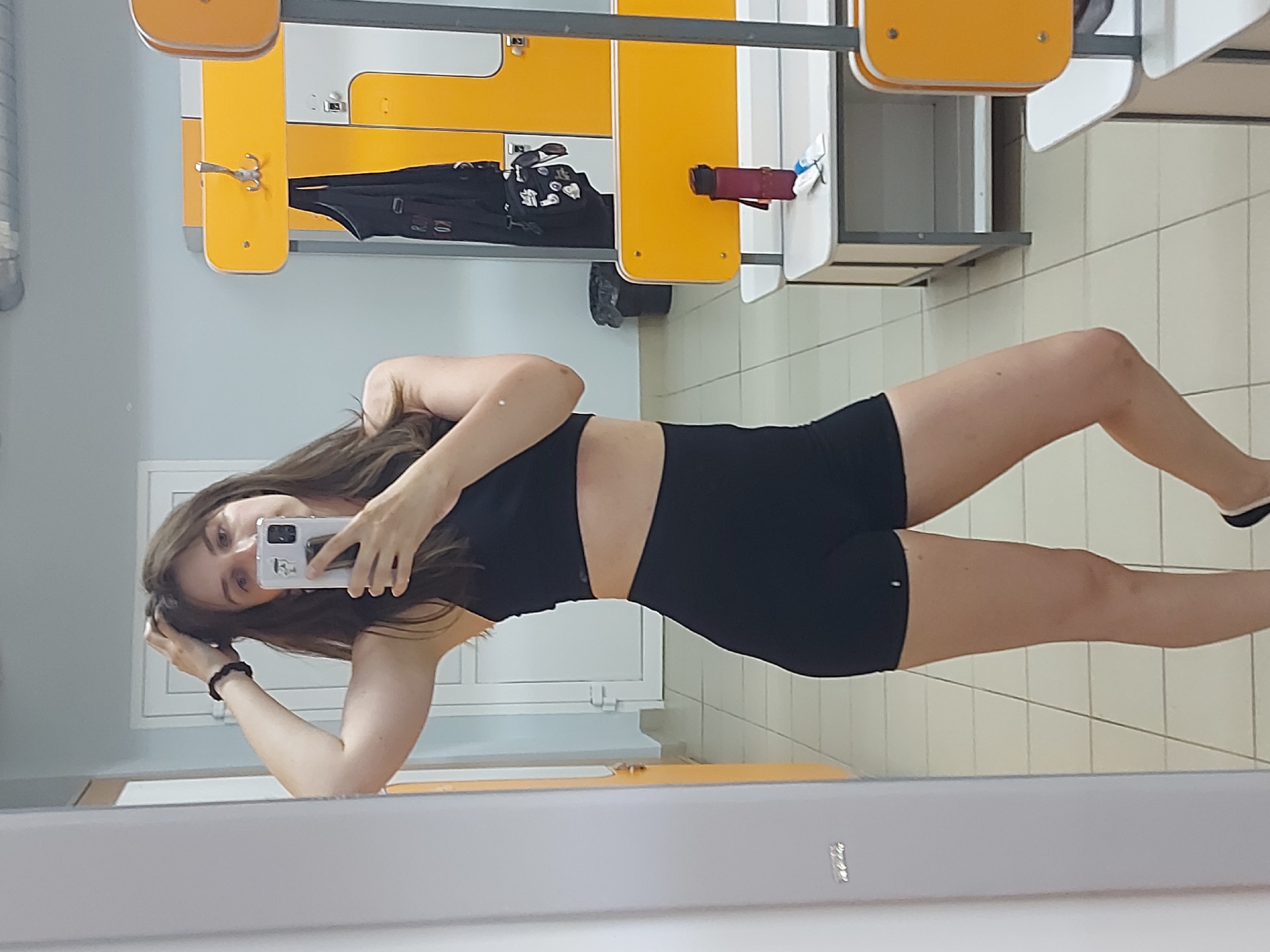 A woman is posing for a selfie in a locker room or changing area. She's holding her phone up with one hand and touching her hair with the other. She is wearing a black sports bra and black shorts. The background includes lockers with yellow and white panels, a closed window with bars, and tiled walls and floors. Based on metadata, the image was taken in Chaikovsky, Russia.