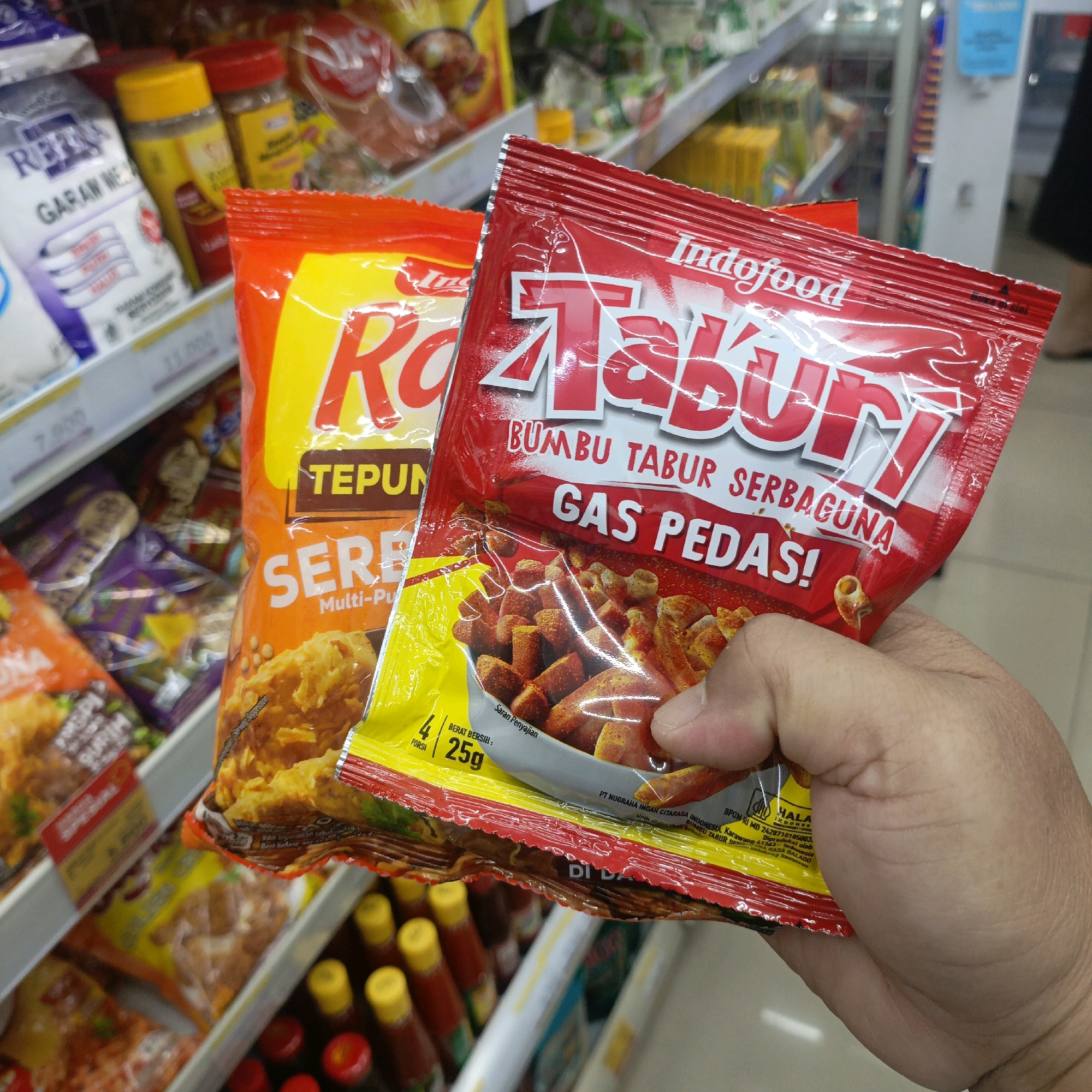 The image shows a close-up of a person's hand holding two packets of Indofood Tabur seasoning in a retail store. The packets are red and yellow, with large text indicating the brand and flavor. The packet in the foreground is red with "Indofood Tabur" printed in large white letters. Below this, it says "BUMBU TABUR SERBAGUNA GAS PEDAS!". The packet behind it is orange and yellow with similar branding. In the background, shelves are stocked with various food products, including bottles with yellow caps and other packaged goods. The scene appears to be inside a modern convenience store or supermarket. The lighting is bright, likely from overhead store lights. The image has a shallow depth of field, with the packets of seasoning in sharp focus and the background items blurred. The specific location within Salatiga, Indonesia, is not determinable from the image itself, as no landmarks or location-specific signage are visible.