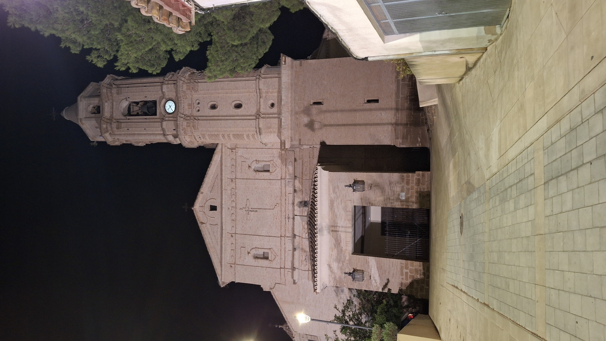The image depicts a nighttime view of the Church of Nuestra Señora de la Asunción in Híjar, Spain. The church is constructed of light-colored brick and features a prominent bell tower. A street paved with interlocking concrete blocks leads towards the church's entrance, which is covered by an awning and flanked by two ornate lanterns. The entrance itself is gated. The sky is dark, indicating nighttime. A tall evergreen tree is visible to the right of the bell tower, partially obscuring the upper portion of the tower. A portion of a building with a tiled roof is visible on the far right. The scene is illuminated by artificial lighting, casting shadows on the brickwork of the church.