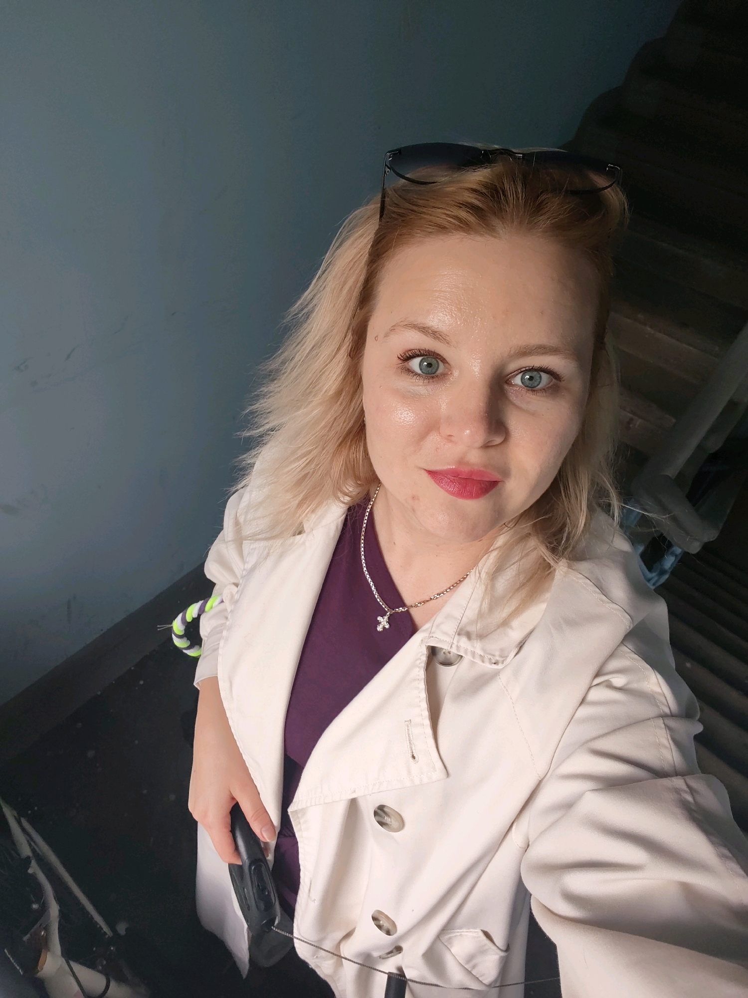 A woman with blonde hair and blue eyes is pictured in a selfie. She is wearing a light-colored trench coat over a purple shirt and a silver necklace with a cross pendant. Sunglasses are perched on her head. Her left hand is visible, holding a black object, possibly a tool or equipment. The background appears to be a dimly lit interior space with a textured wall and wooden stairs. Based on the provided metadata, this image was taken in Izhevsk, Russia.
