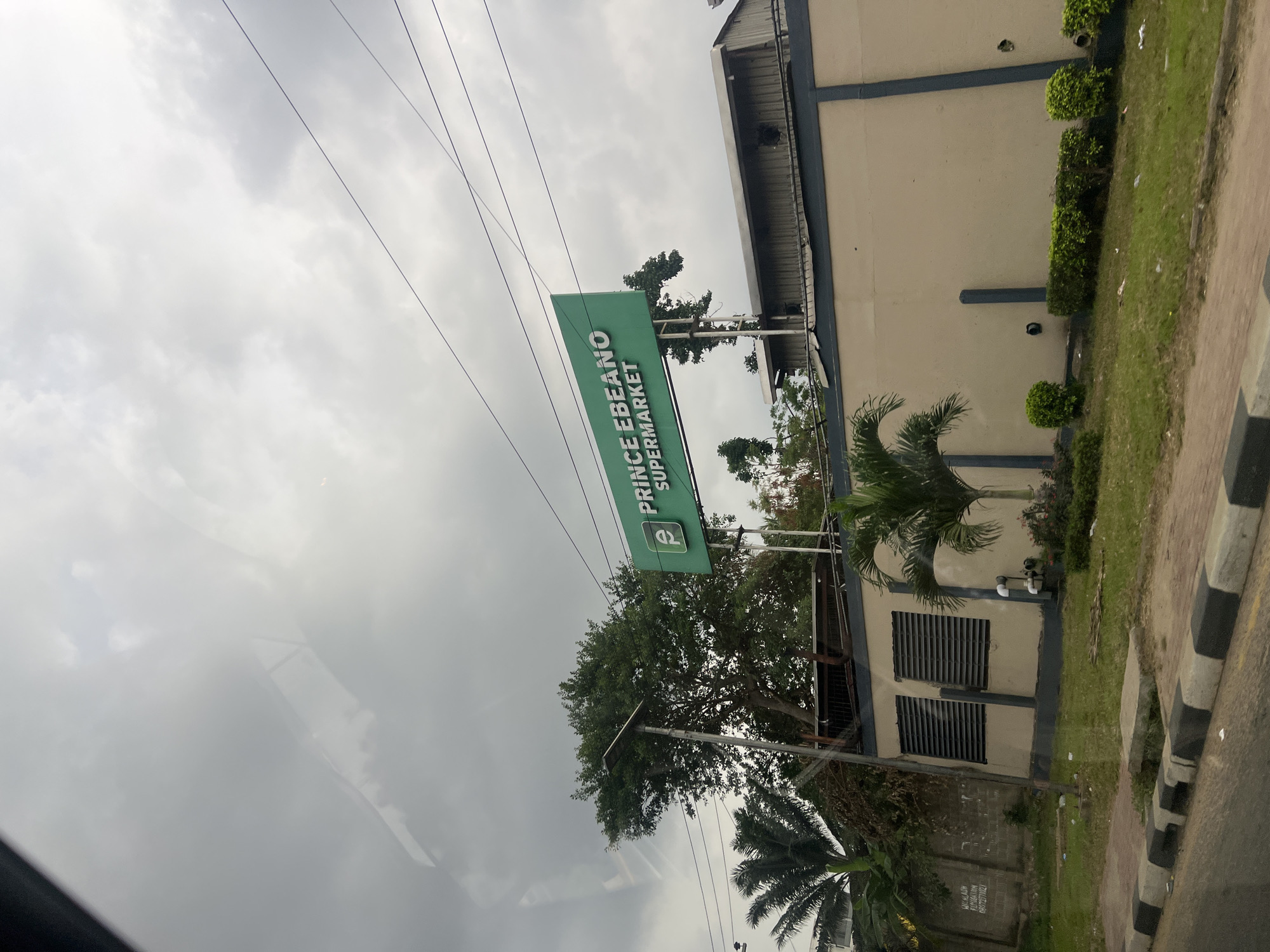 The image shows the Prince Ebeano Supermarket in Ikeja, Nigeria. The supermarket sign is visible in green with white lettering. The building is beige with dark blue trim. There is a sidewalk with black and white striped curbs. Trees and other vegetation are also visible. The sky is overcast. Power lines run across the top of the image.