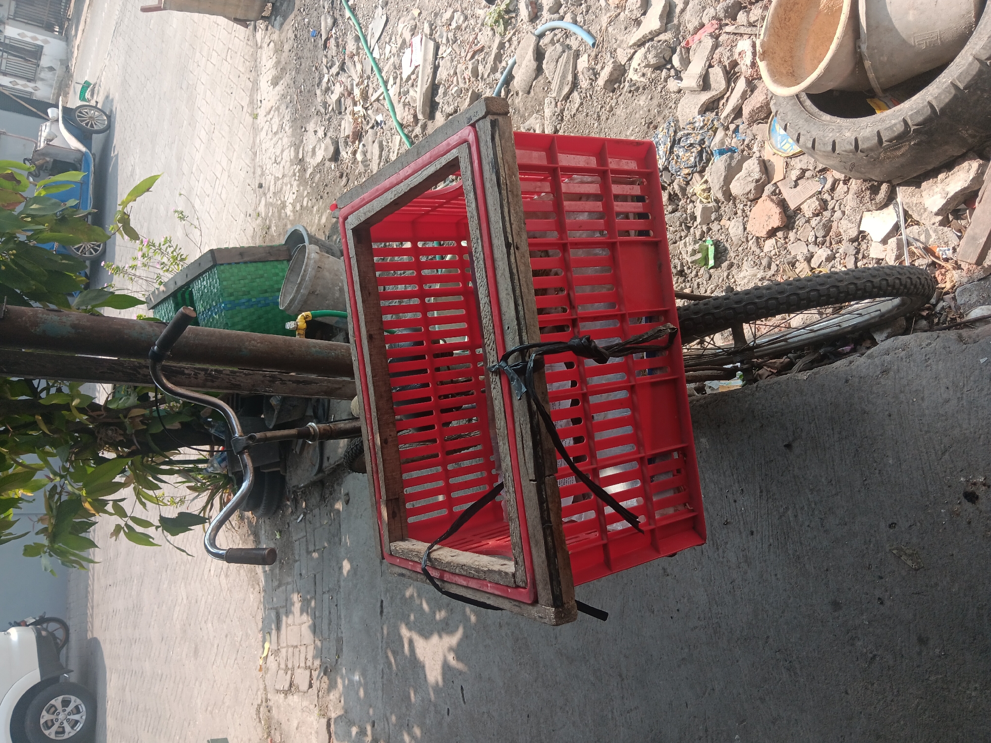 The image captures an outdoor scene in Batursari, Indonesia, with a focus on a bicycle equipped with a large red plastic crate. The bicycle is parked on a paved surface, likely a sidewalk, adjacent to a dirt and debris-strewn area. The red crate, reinforced with wooden planks around its rim, is securely fastened to the bicycle with black straps.

In the background, a white car is partially visible on the left. Further back, a blue and white vintage-style car is parked on a street paved with interlocking bricks. Green foliage from a tree shades the area, and a green woven basket and a grey bucket are near the tree. Scattered on the ground are various pieces of rubble, stones, and what appears to be discarded tires. The overall environment suggests a somewhat utilitarian or informal setting. There are no people clearly visible in the image. The lighting indicates it is daytime with sunlight casting shadows.