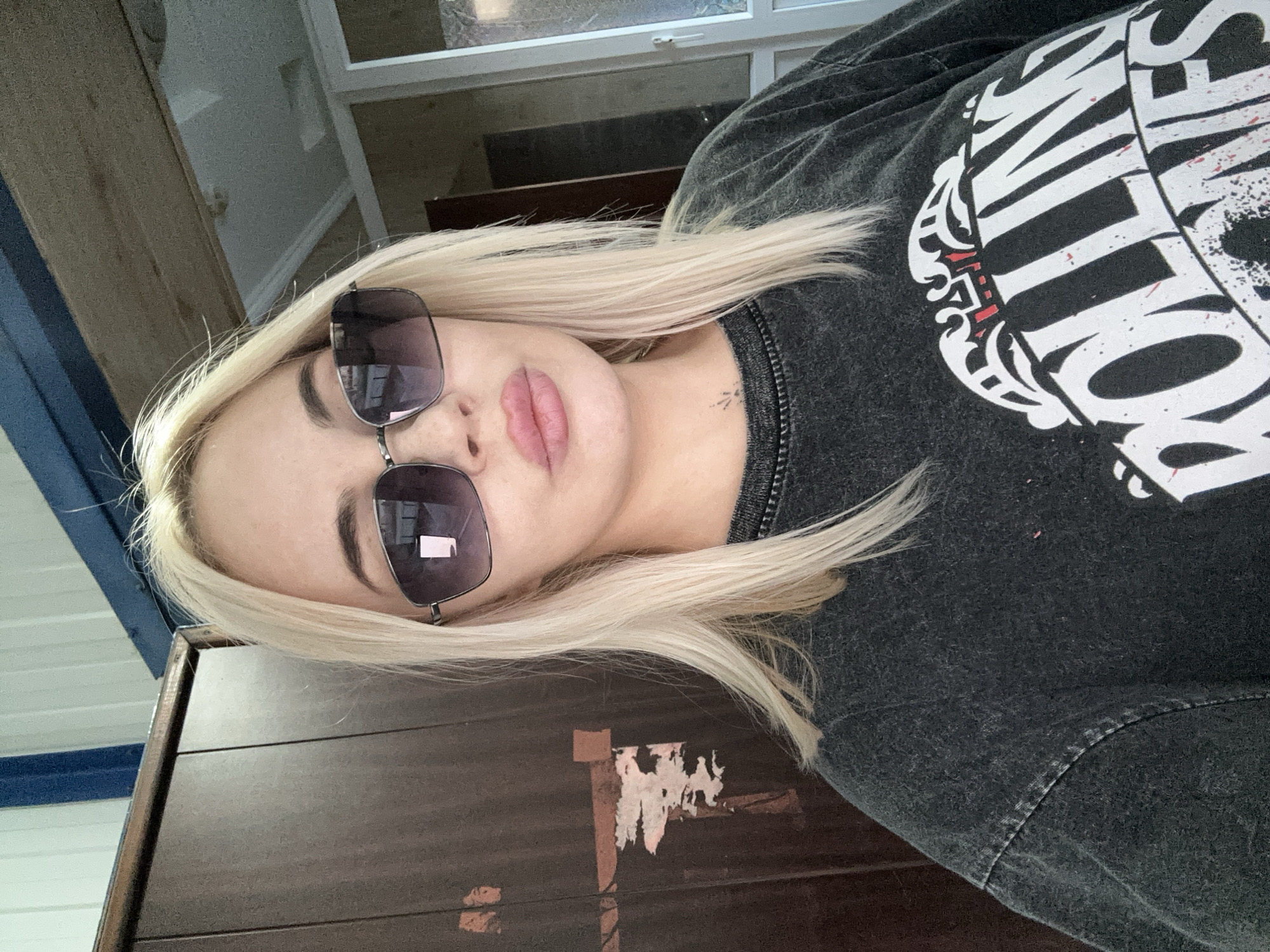 A fair-skinned woman with blonde hair is wearing sunglasses and a gray t-shirt. She is lying down on a dark brown wooden surface. The woman has a small tattoo on her neck. The background includes a white wall and a glimpse of stairs. The location is Высокий, Russia.