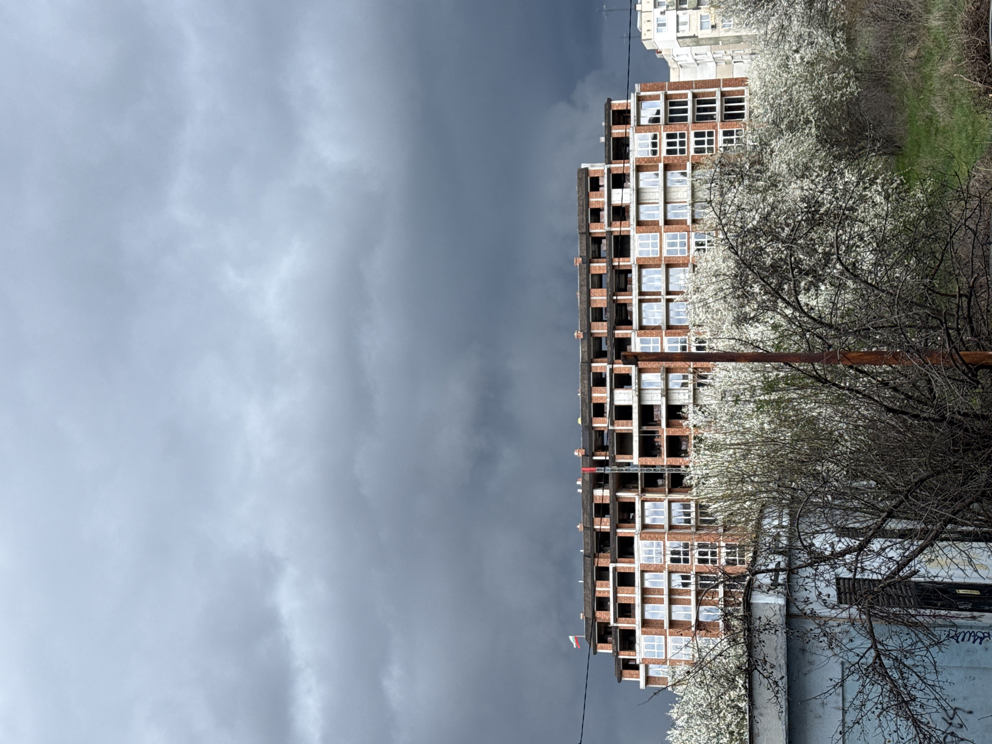 An eye-level, vertical, and full shot depicts a building under construction in Sofia, Bulgaria. A gray cloudy sky dominates the scene. A multi-story building in the center of the frame is primarily brick, with many window openings; some have white frames installed. A red crane stands atop the building. White blossoming trees fill the right side of the shot. An adjacent existing white apartment building is visible on the right. A building is at the bottom right corner. Powerlines are visible. No people are visible.