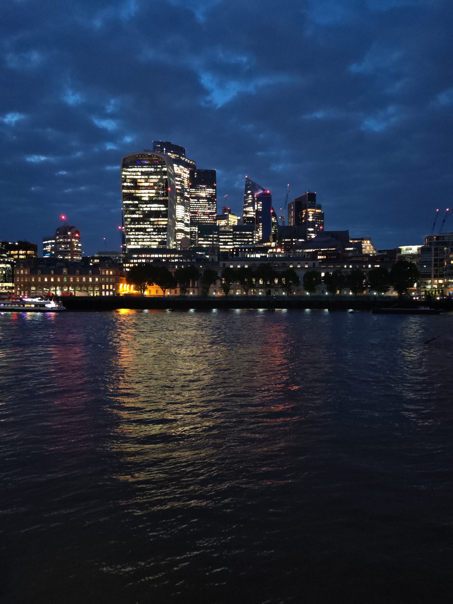This is a nighttime shot of the City of London, UK, taken from across the River Thames. Several modern skyscrapers are visible, their windows brightly lit. The "Walkie Talkie" building is prominently featured. The river is dark, reflecting the lights of the buildings and passing boats. The sky is a deep blue with scattered clouds. Trees and other buildings line the riverbank. A single passenger boat is visible on the water.