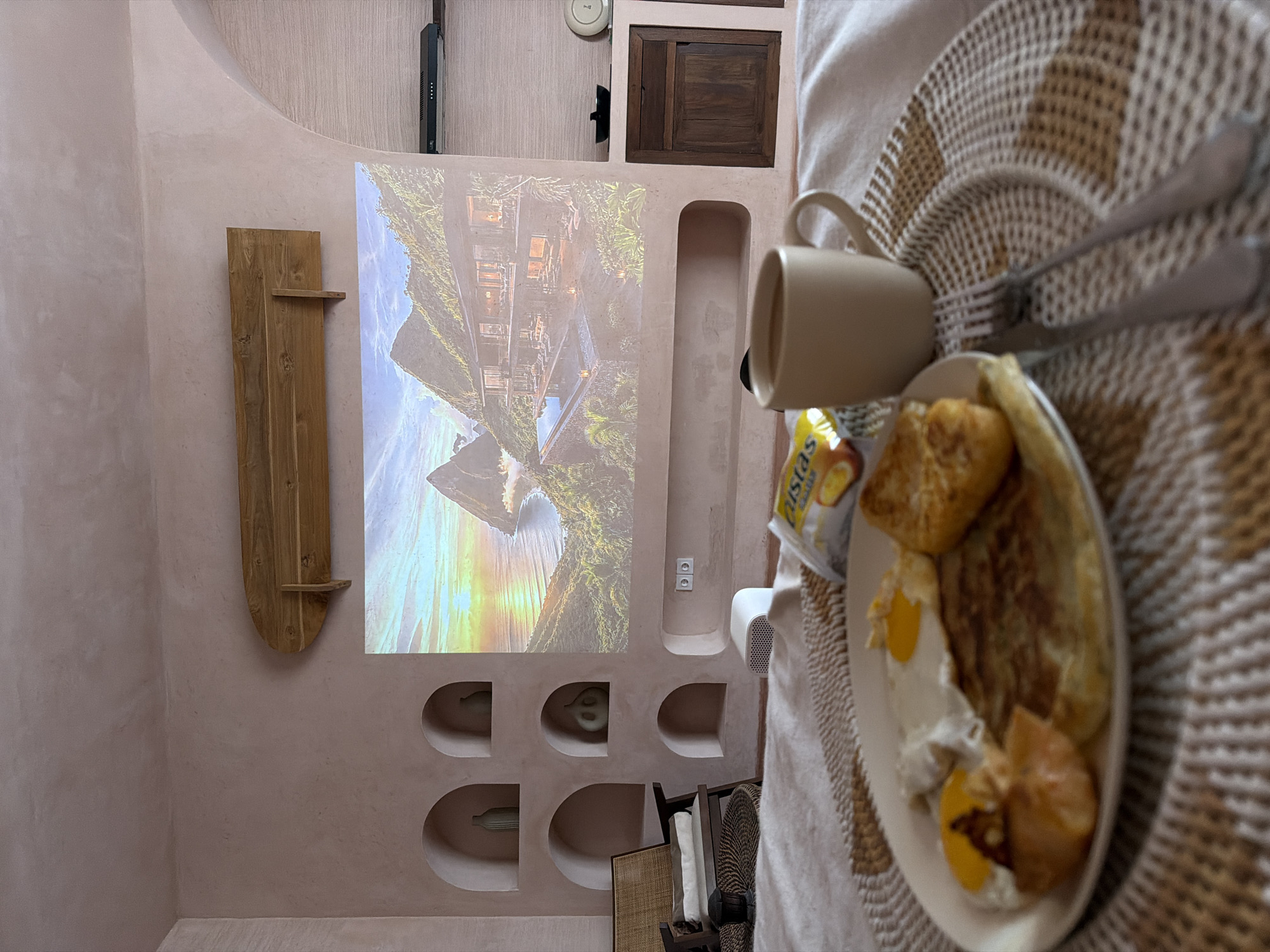 A breakfast plate and coffee cup sit on a placemat on what appears to be a bed in an interior room. The breakfast plate contains eggs, pastries, and a packaged food product. A fork and knife are placed on top of the placemat.
In the background, a screen displays a landscape image with trees and water, seemingly projected onto the wall. The room's wall features decorative niches with small objects and wooden furniture. There is a television on the wall, along with electrical outlets. The room has a pinkish hue and appears to be in North Kuta, Indonesia.