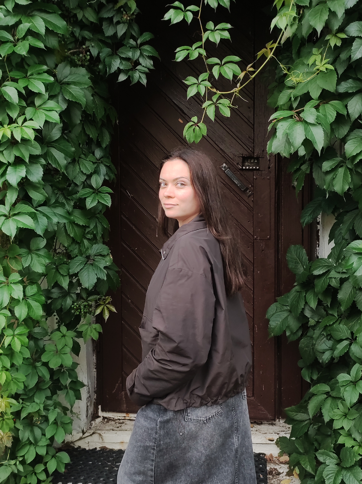 A young woman with long brown hair stands to the left of a dark brown wooden door, partially obscured by lush green ivy. She is wearing a dark brown bomber jacket and a grey denim skirt. The woman is looking over her left shoulder towards the camera with a slight smile. The door has visible wood grain and what appears to be a metal latch. The ivy is dense and leafy, framing the doorway. The ground in front of the door appears to be paved. There are no other people or animals visible in the image. This scene is consistent with an entryway to a building in Saint Petersburg, Russia, as such wooden doors and abundant greenery are common architectural and horticultural features found throughout the city, particularly in older residential areas or dacha properties.