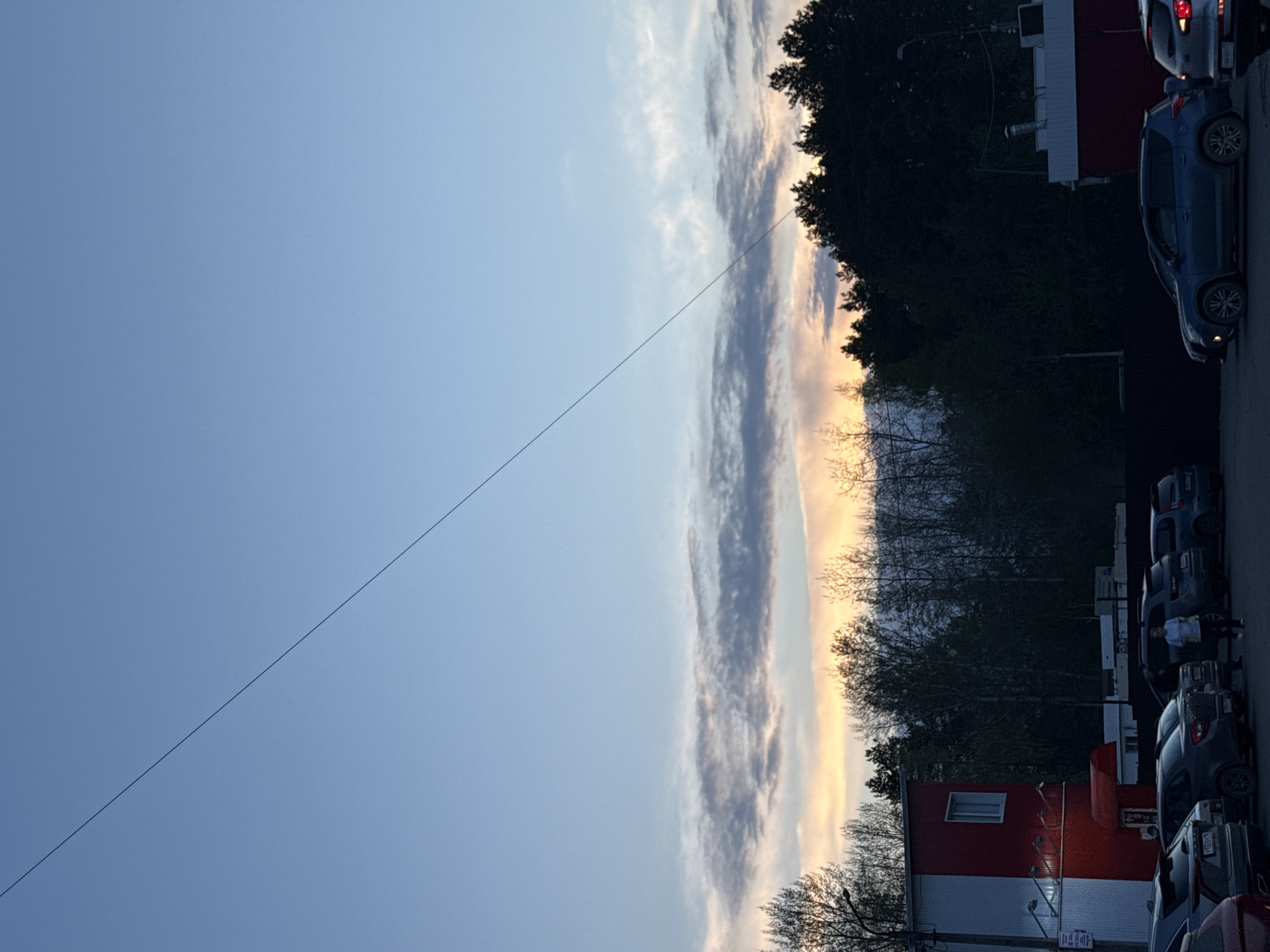 The image captures a street scene in Chaikovsky, Russia, during what appears to be either dawn or dusk. The sky transitions from a clear blue to a band of clouds near the horizon, tinged with yellow light. A power line stretches across the sky.
On the right side of the frame, several parked cars line the street. A two-story building with a red upper level and a white lower level is visible amidst the trees. The trees are mostly bare, suggesting it might be early spring or late fall. There's also a dark building. There are no people in the picture.