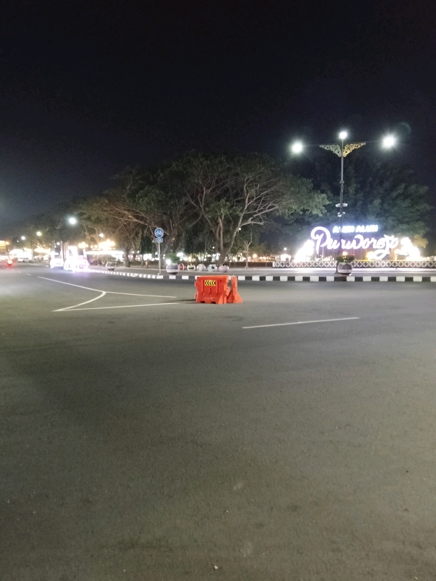 The image captures a nighttime view of a street intersection in Purworejo, Indonesia. A large, illuminated sign spelling out "AHMAD ALUN PURWOREJO" dominates the background, with decorative streetlights in the mid-ground. The foreground shows a paved road with white lane markings. An orange traffic barrier with reflective tape is positioned on the right side of the road. Trees line the street behind the intersection. A few vehicles are visible in the distance, their headlights illuminating the scene. The overall atmosphere is that of a well-lit urban space at night.