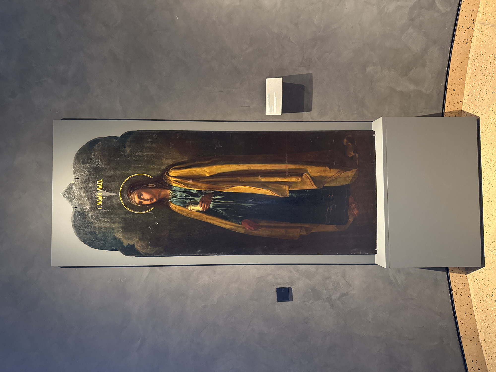 This is a photograph of an icon depicting Saint Mary Magdalene, displayed in an art museum. The icon is framed by a dark arch and features a halo around the saint's head. She is wearing a blue garment and a golden cloak, and holding a vessel in her right hand. The background is dark, with swirling textures suggesting clouds or a mystical atmosphere. Golden Cyrillic text is visible above the saint's head. The icon is mounted on a white pedestal and stands against a grey, textured wall. A small informational plaque is affixed to the wall to the right of the icon. The floor is a terrazzo material. The overall lighting suggests an indoor exhibition space. This icon is part of the collection of the Perm State Art Gallery in Perm, Russia.