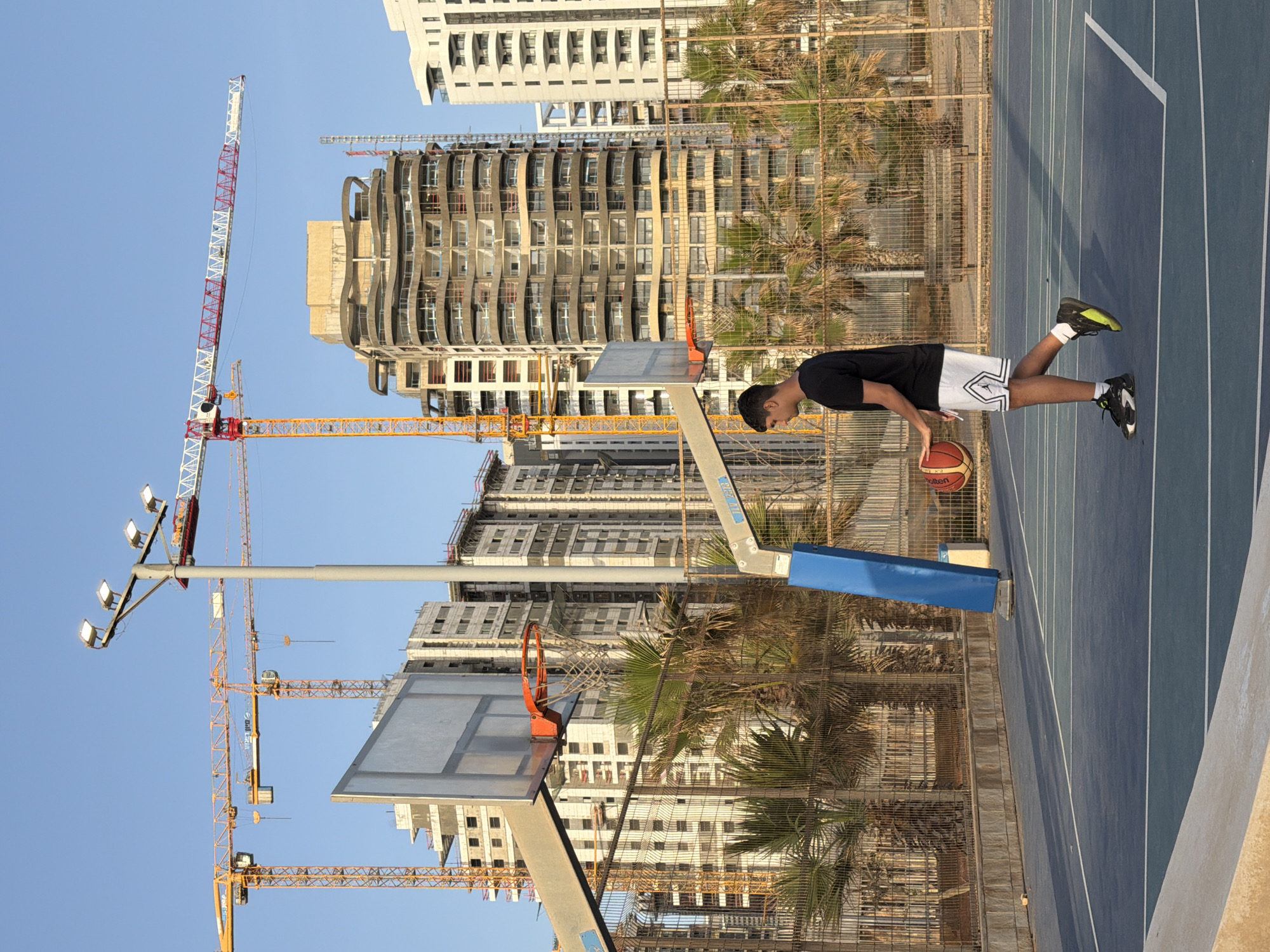 A male basketball player practices on a blue court with basketball hoops, amidst construction sites in Rishon LeZion, Israel. Construction cranes and modern buildings form the backdrop. The player is wearing black t-shirt and patterned shorts. He is dribbling a basketball. The court is surrounded by a wire fence and palm trees.