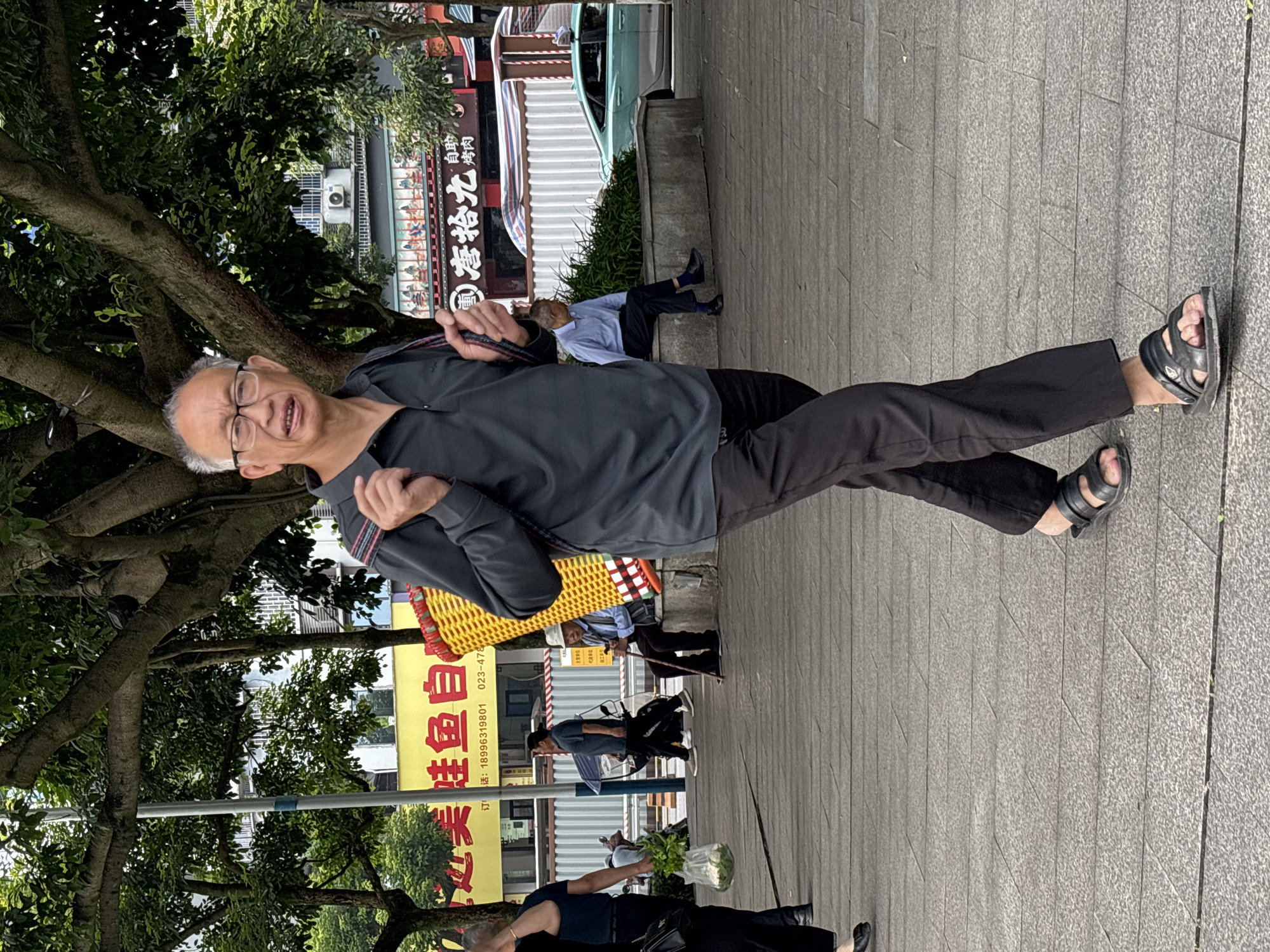 An outdoor, eye-level, full-shot photograph captures an elderly East Asian man walking on a paved walkway in Jiangjin, China. The man, wearing glasses, a dark grey polo shirt, and black trousers, carries a large, yellow woven basket on his back. He is walking towards the viewer. The walkway is made of grey rectangular pavers. Behind him, a large tree with broad branches and green leaves provides shade. In the background, there are buildings with signage in Chinese, including one with red characters on a yellow background. A teal-colored car is visible on the right side of the image. Other pedestrians are present in the mid-ground, some sitting and some walking. The overall lighting suggests a daytime scene with diffused sunlight.