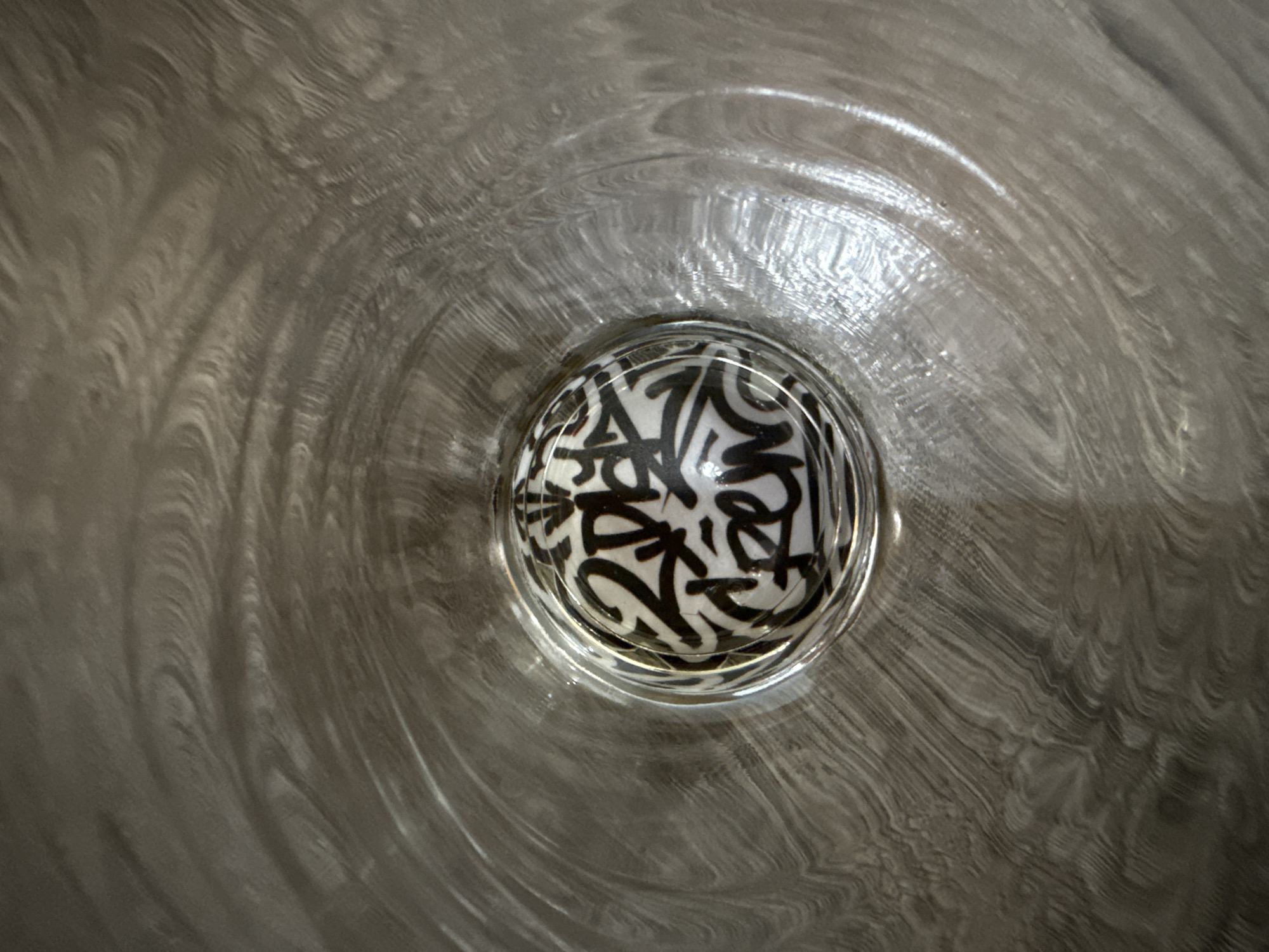 The image displays an overhead perspective into a metallic, cylindrical basin or container located in Madrid, Colombia. The inner surface of the basin exhibits a textured pattern of concentric and swirling brush marks or grooves. At the bottom center of the basin, a small, white spherical object is partially submerged in a shallow layer of clear liquid. The sphere is densely covered with black, graffiti-style script and stylized lettering. Identifiable elements within the script include characters resembling "2DES" and "CREW". No people are visible in the scene.