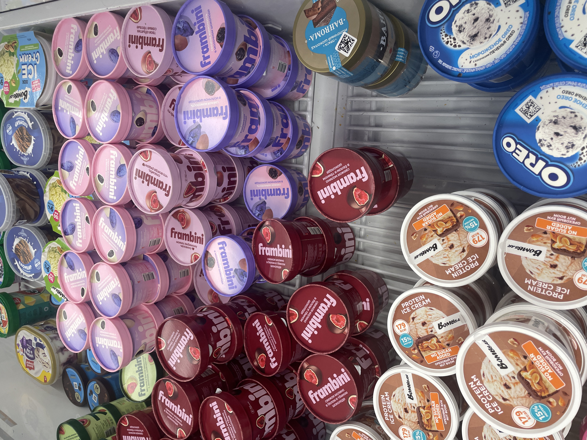 The image shows a refrigerated display case filled with various brands and flavors of ice cream. Dominating the scene are numerous containers of "Frambini" ice cream in different flavors like raspberry in dark chocolate, plum, and honeysuckle in milk chocolate, cascading from top left to bottom right. Alongside, are tubs of "Bombbar" protein ice cream with nut flavors, and "Oreo" cookie ice cream. Other brands, like "Bahroma" and "Plomber", are also visible. The display suggests a retail setting, likely a supermarket or convenience store in Izhevsk, Russia, as indicated by the Russian text on the product labels. The arrangement implies an intentional, possibly promotional, layout of the ice cream selection.