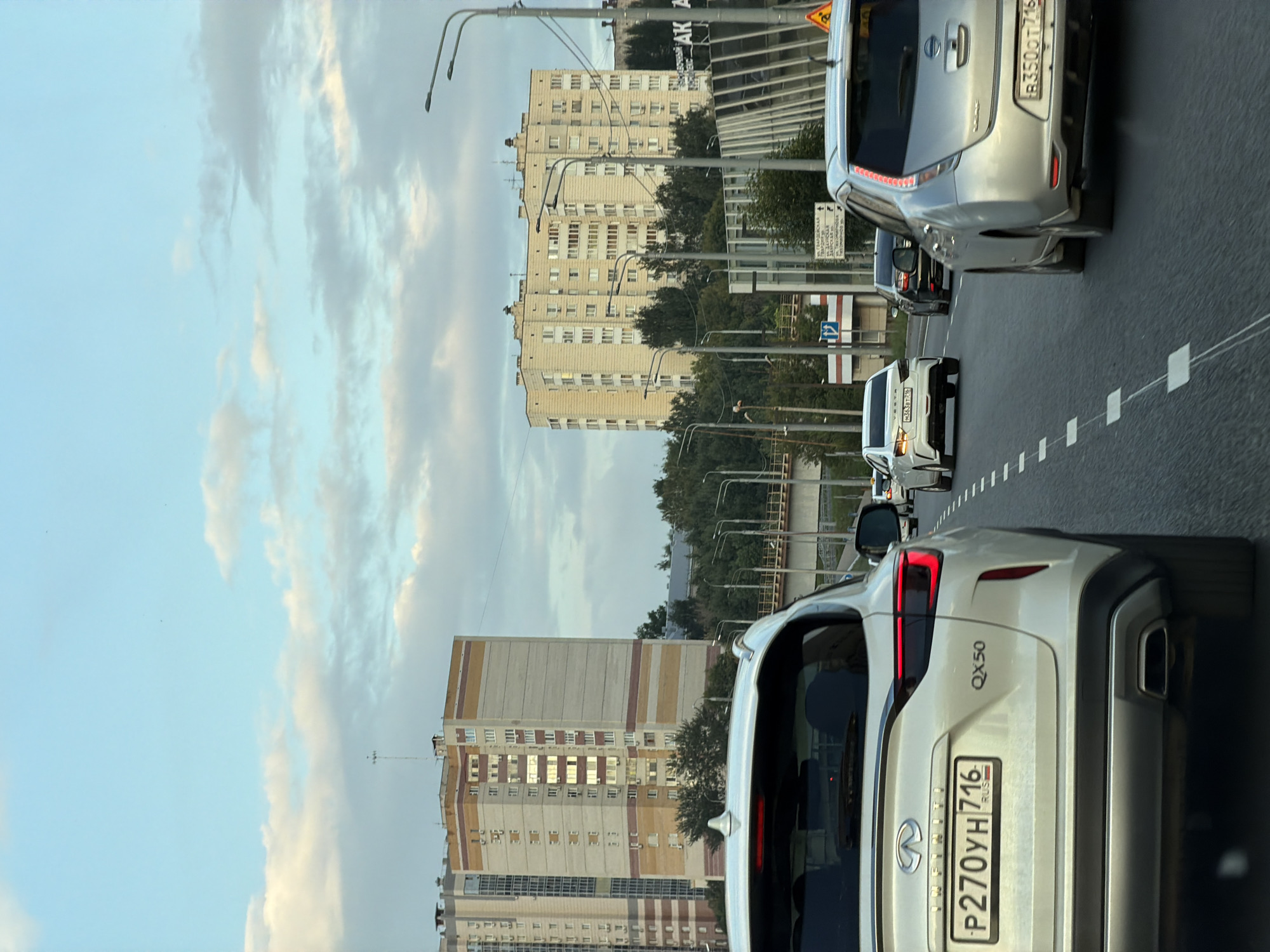 The image shows a view from inside a white SUV driving on a multi-lane road in an urban environment. In the foreground, the rear of the white SUV is visible, displaying a license plate with the inscription "P270YH 716 RUS". Other vehicles are present on the road, including a silver Nissan Leaf with license plate "B3500T 716" and a white sedan with license plate "M363BT 716". In the background, two tall, multi-story apartment buildings are visible, one on the left and another on the right. The sky is partly cloudy. Streetlights and some trees are also present. The scene suggests a daytime, possibly late afternoon, setting.
