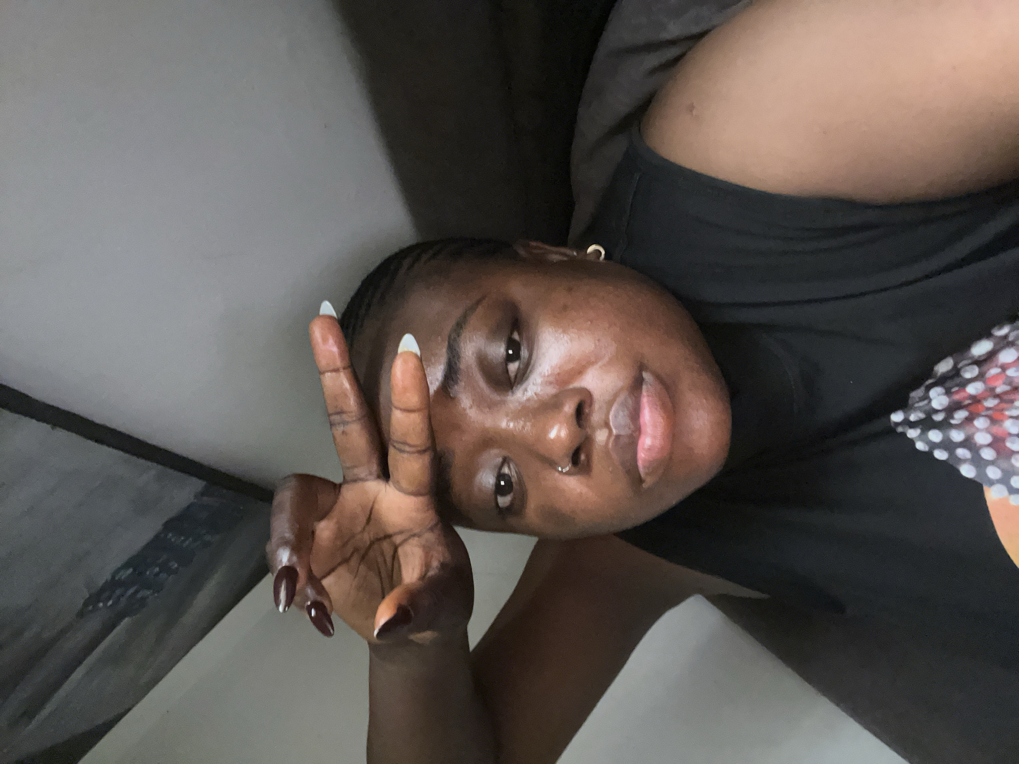 In Abuja, Nigeria, a close-up, eye-level selfie depicts a young Black woman. She is positioned in the lower right of the frame, with her head tilted slightly forward and to the right. Her right hand is raised to her forehead, with her fingers splayed, as if to shield her eyes from the sun or indicate weariness. She wears a black, sleeveless top. Her hair is styled in cornrows. She has a nose ring and a hoop earring in her left ear. The background is dimly lit and appears to be an indoor environment, possibly a bedroom, with a large, angled surface visible in the upper left quadrant, potentially part of a wall or furniture. The lighting suggests it is either late evening or early morning.