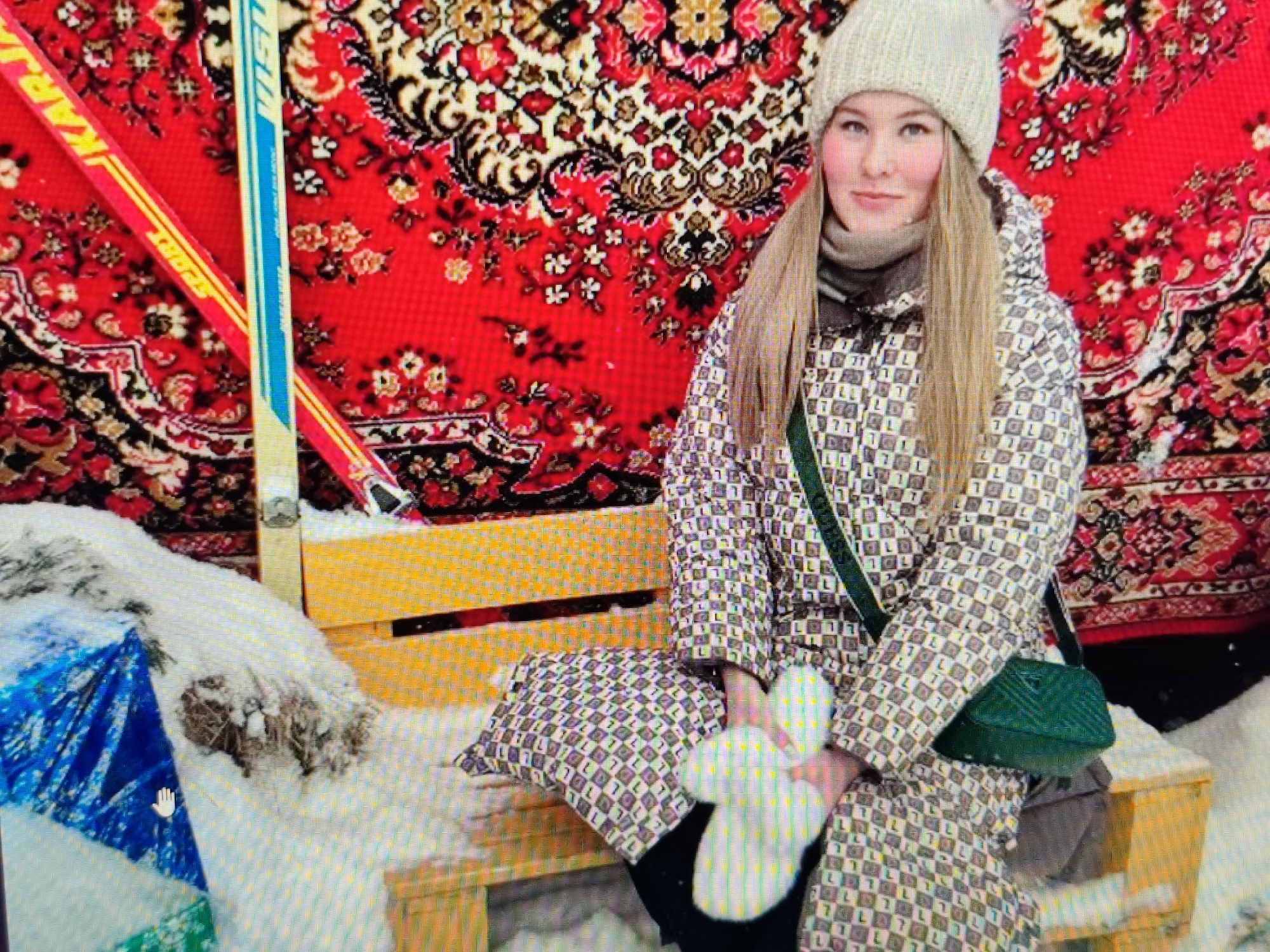 A woman in a patterned winter coat and a knitted hat sits on a wooden bench. She is holding a white object in her hands. Behind her, a red rug with a floral pattern is hung on a wall. To her left, two ski poles are propped up against the rug. The ground is covered in snow, with a blue tarp and some branches visible on the left side of the image. The overall scene suggests a cold, winter environment. The specific location in Syktyvkar, Russia is not discernible from the image alone.