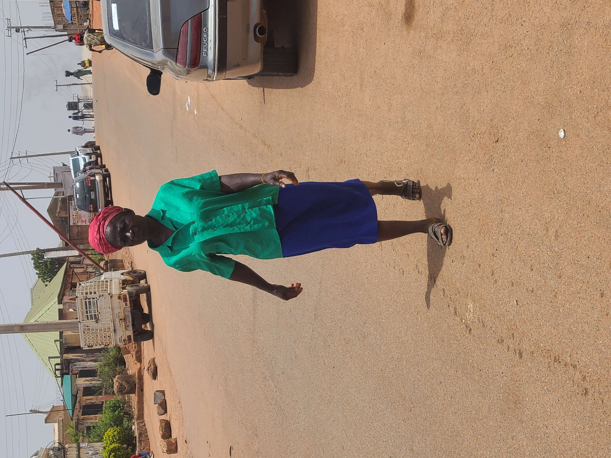 An elderly woman is standing on a brown, paved road in what appears to be a residential area. She is wearing a green button-down shirt, a blue skirt, a red headscarf, and brown sandals. A silver Peugeot car is parked partially on the road beside her. In the background, there are other vehicles, buildings, and utility poles. There are people walking on the street in the distance. Based on the appearance of the buildings, vehicles, and the general environment, it can be inferred that this image was taken in the city of Jos, Nigeria.