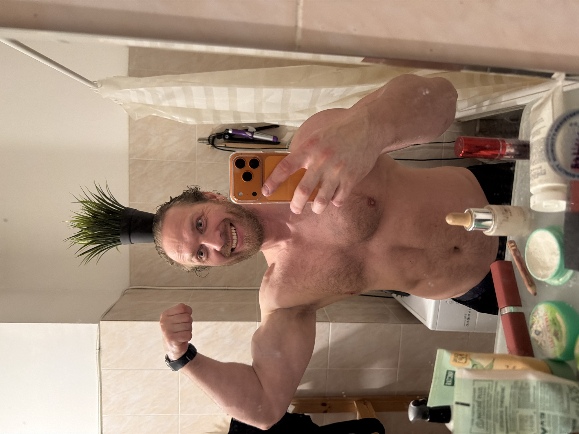 An adult male is captured in a mirror selfie within a bathroom setting, purportedly in Saint Petersburg, Russia. The subject, shirtless, possesses a muscular build and a light beard, smiling broadly at his reflection. His left arm is flexed, showcasing a bicep, and a dark smartwatch is visible on his wrist. He holds an orange smartphone with his right hand, likely operating the camera. A small artificial green plant in a black pot is balanced on top of his head. The background, visible in the mirror, features beige tiled walls, a white washing machine, and a partially drawn white striped shower curtain on a metal rod. Hair styling tools are suspended from a hook on the wall behind the man's right shoulder. The foreground displays a collection of personal care products and cosmetics on the vanity surface, including tubes, bottles, and various containers.