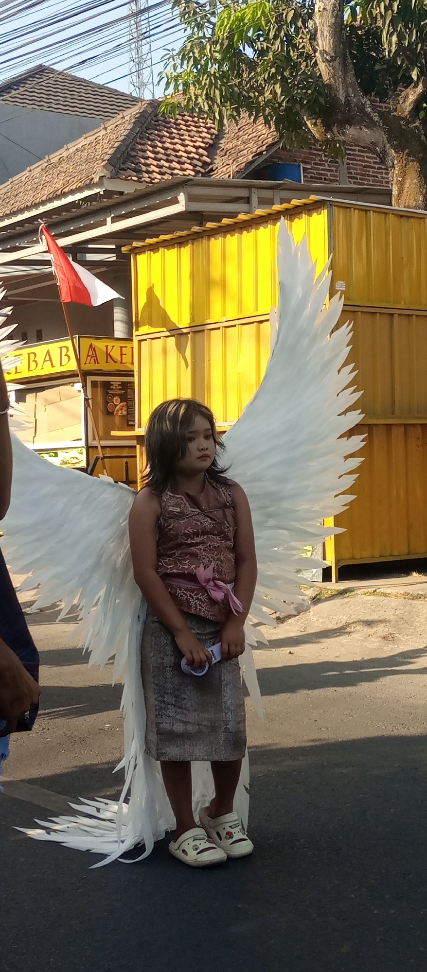 A young girl with dark, layered hair and bangs stands in the middle of a paved street, wearing a costume resembling angel wings. The wings are large and white, made of layered white material. She is also wearing a brown patterned sleeveless top with a pink sash tied around her waist, and a gray patterned skirt that extends to her knees. On her feet, she wears white slip-on shoes with colorful designs. She holds a small white object in her right hand.
In the background, there are several structures. To the left, a red and white Indonesian flag is attached to a pole. Behind that, a building with a tiled roof and yellow siding is visible, with a sign that reads "KEBAB AK..." partially visible. The ground around the girl is paved and appears to be a street or walkway, with shadows cast by the sunlight. Trees with green leaves are also visible in the background, along with electrical wires strung overhead. The overall scene suggests an outdoor event or parade. The location is identified as Jekulo, Indonesia.