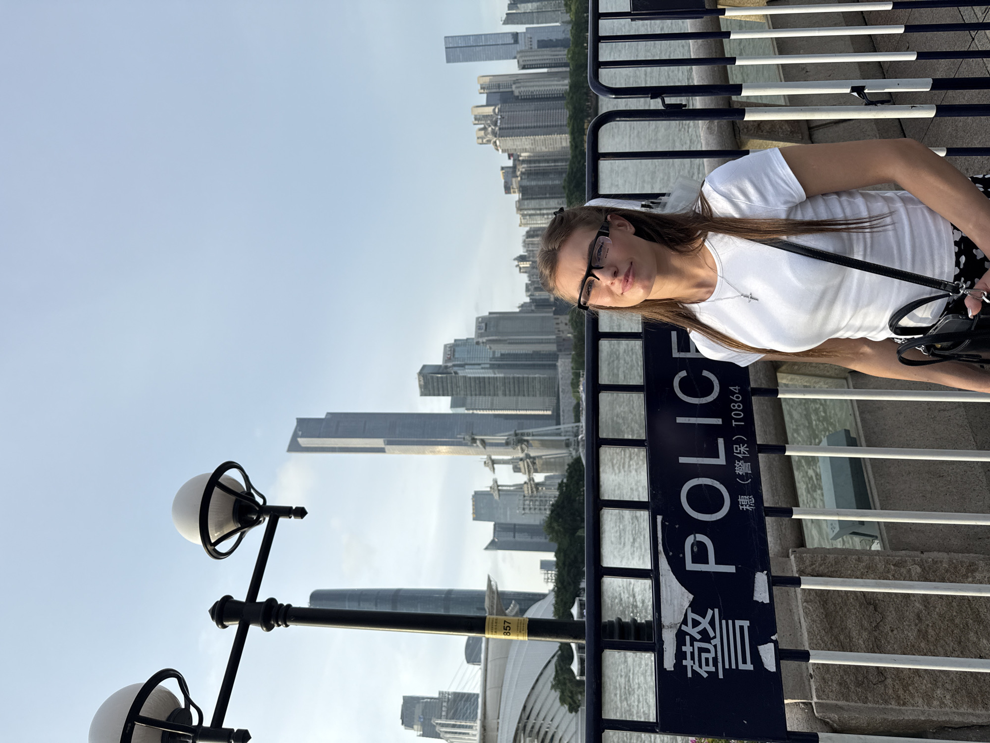 A young Caucasian woman with long brown hair and glasses is standing in front of a city skyline. She is wearing a white t-shirt and a black and white patterned skirt. She is holding a black purse on her shoulder. Behind her, there is a body of water and a fence with a "POLICE" sign in English and Chinese. The skyline features tall buildings, including skyscrapers. The scene appears to be in Haizhu District, China, based on the presence of Chinese text on the police sign and the architectural style of the city. The sky is blue with some clouds. The lighting suggests it is daytime, possibly late afternoon.