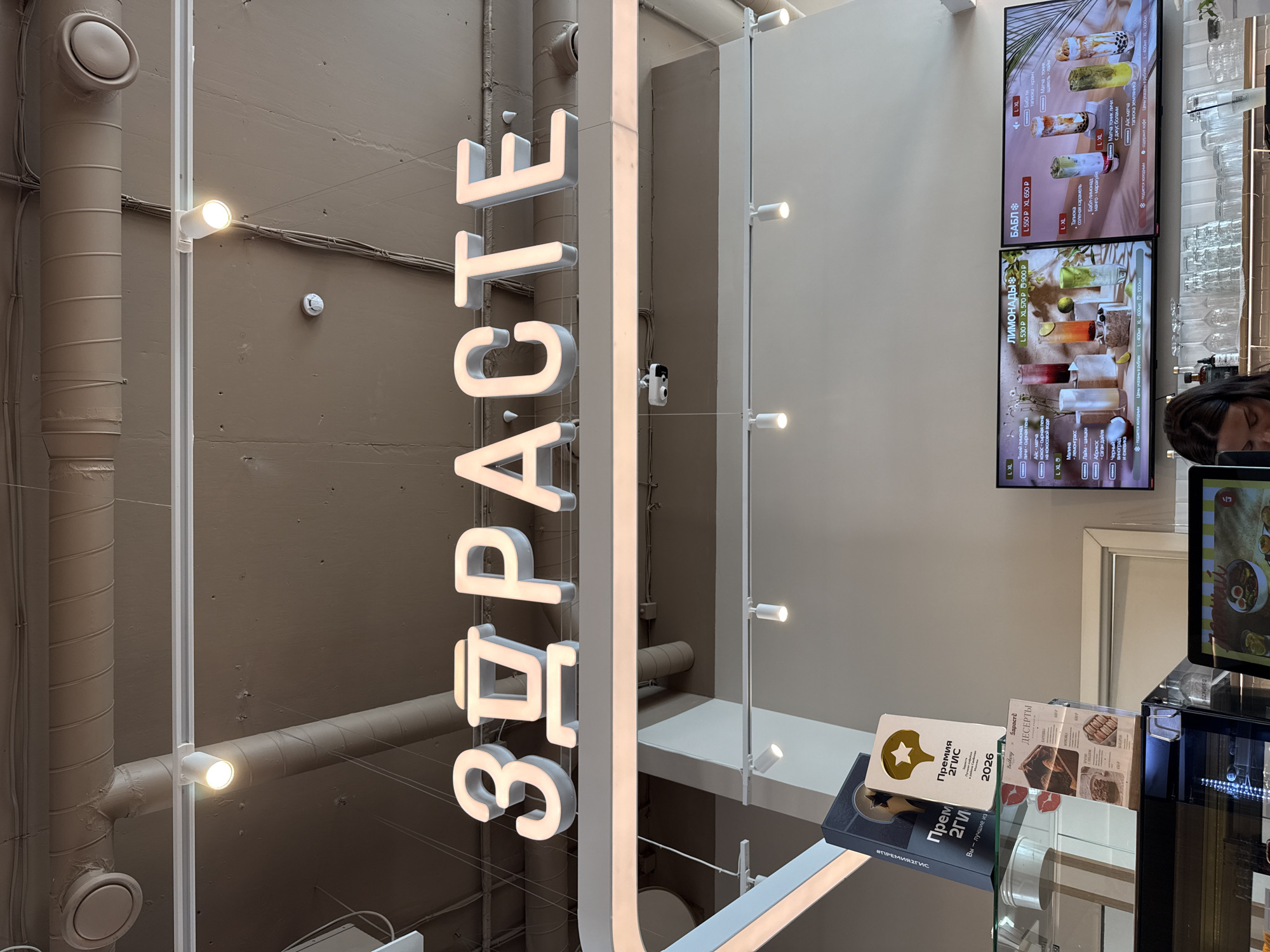 This is a medium-angle, eye-level interior shot of a cafe or restaurant. A large, illuminated sign with white, blocky Russian letters spelling "ЗДРАСТЕ" (ZDRASTYE), meaning "Hello", is prominently displayed overhead. Below the sign, a menu board is visible, displaying drinks categorized as "ЛИМОНАДЫ" (Lemonades) and "БАБЛ" (Bubble Tea). The menu board shows images of colorful beverages and their prices in rubles. To the left of the menu board, a stack of awards or certificates is visible, one featuring a golden star and the text "ПРЕМИЯ 2ГИС" (2GIS AWARD) with the year "2026". In the foreground, a counter is visible with glassware and a woman's head and shoulders in the lower right corner, seemingly an employee. The environment appears modern and clean, with exposed ceiling pipes and track lighting contributing to the industrial-chic aesthetic. The lighting is bright, suggesting it's daytime or well-lit indoors. The overall impression is that of a contemporary establishment focused on beverages and a welcoming atmosphere. The presence of the "2GIS AWARD" suggests the establishment is likely located in Russia, and specifically in Moscow, as 2GIS is a popular Russian directory and navigation service.