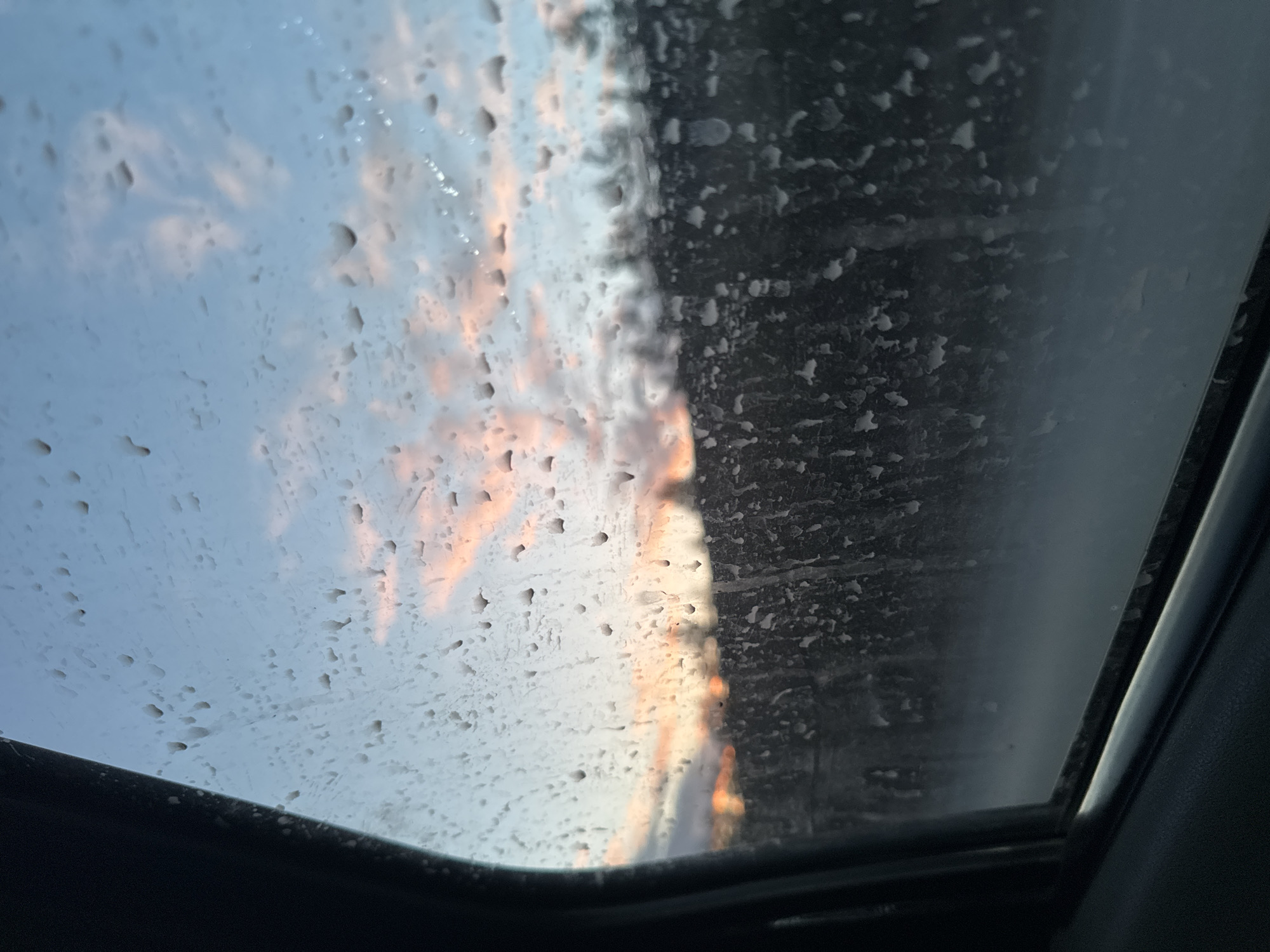 The image shows a view through a sunroof, partially obscured by water droplets. Outside, there is a glimpse of a sky with clouds tinged with orange and light blue. The sky is on the left of the view and to the right there is a dark grey area covered in water droplets. The edge of the sunroof frame is visible at the bottom and sides of the image, appearing black. The image was presumably taken in Iiul'skoe, Russia.