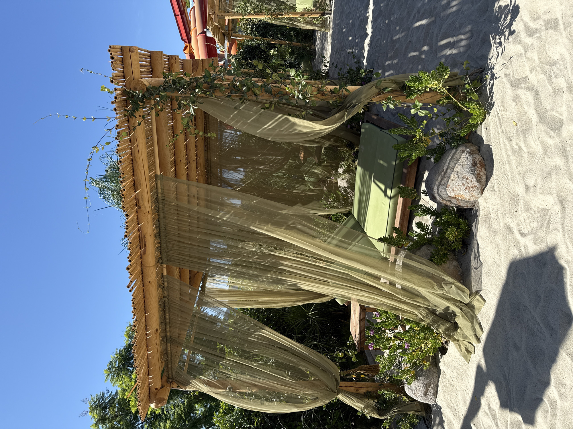 This is a photograph of a bamboo-roofed cabana with light green sheer curtains and a green mattress, located on a sandy beach. The cabana is decorated with climbing plants and surrounded by rocks and small green plants. In the background, there are trees, a portion of a red water slide, and another similar cabana. The scene appears to be a resort area in Bodrum, Turkey, suggesting a sunny day with clear blue skies. There are no people visible in the image. The shadows cast on the sand indicate direct sunlight.