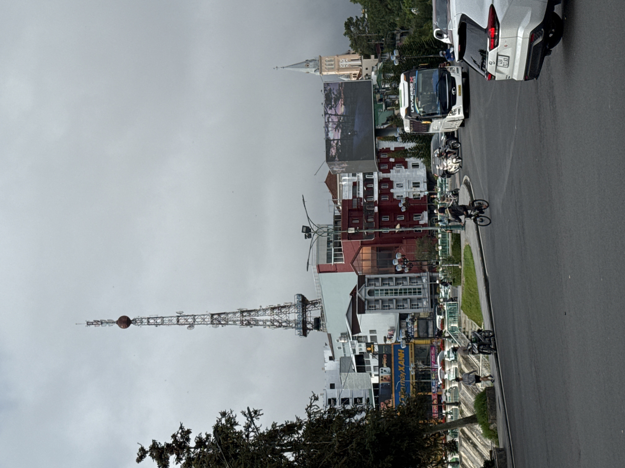 The image captures an urban street scene in Da Lat, Vietnam, under an overcast sky. The perspective is from a slightly elevated position, looking across a road towards a mix of architectural styles and city activity.
On the left side of the frame, a tall, lattice-structured communications tower with a distinctive circular dish near its peak dominates the skyline. In the mid-ground and extending into the background, various multi-story buildings line the street, featuring diverse designs with prominent red and white facades. Further in the distance, a prominent church with a light-colored steeple adds a historical and religious landmark to the cityscape. Several large billboards are integrated into the urban fabric, one of which displays a scenic image of a sunset or nature.
The street is active with traffic and pedestrians. A white SUV is parked on the far right, displaying a license plate "49A - 7X7.63". Behind it, a white bus with "UNIVERSE X" emblazoned on its side is visible. A person on a bicycle cycles along the road, and multiple motorcycles are present, either parked or in motion. Along the sidewalks and medians, greenery from trees and grassy areas breaks up the concrete environment.
The weather appears to be cloudy and overcast, without direct sunlight, suggesting a cool or gloomy daytime. Visible text on banners and signs includes partial Vietnamese text such as "...máy XANH" (likely "The Gioi Di Dong/Dien May Xanh" - a major electronics retailer) and "P R O", further confirming the Vietnamese setting.