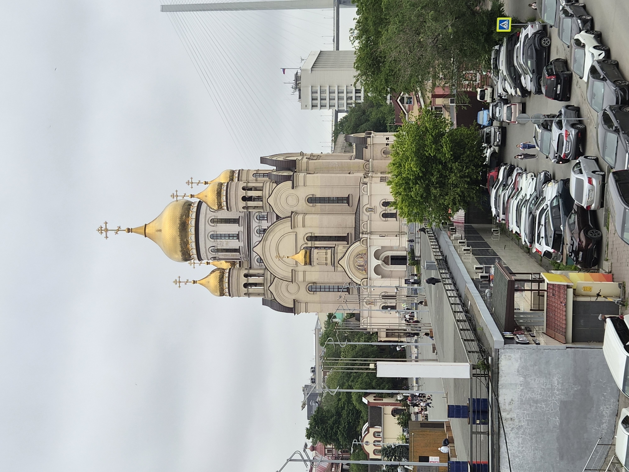 The image shows a view of the Church of the Archangel Michael in Vladivostok, Russia. The church, with its distinctive golden domes, is the central subject of the photograph. The scene is overcast, with a cloudy sky dominating the upper portion of the image.
In the foreground, a multi-level parking lot is visible, filled with numerous cars. Some people are walking or standing in the parking lot and on a raised walkway above it. To the left of the church, there are some smaller buildings and trees. In the background, a large suspension bridge can be seen spanning across the water, a prominent landmark of Vladivostok. A tall white building with a Russian flag on top is also visible near the bridge. The overall environment suggests an urban setting, likely a public space with both religious and infrastructure elements.