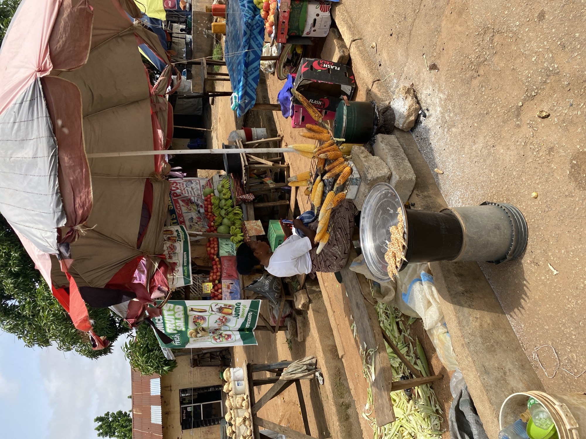 This is a photograph taken outdoors, likely in a market setting in Bukuru, Nigeria. The image is oriented vertically, with the top of the scene appearing on the left.
The primary subject is a woman, dressed in a white short-sleeved top and a patterned skirt, sitting at a stall. She appears to be attending to her goods, which include numerous ears of corn, some of which are displayed on a rack. In front of her, there is a large black container with a clear lid, filled with corn kernels.
The stall itself is covered by a large, multi-colored umbrella, providing shade. Behind the woman and the umbrella, there are various items for sale, including a display of fruits like bananas and tomatoes, and some crates. Signs are visible, one of which reads "fresh FRUITS SALAD" with images of fruit dishes, and another with "Intact: 01131225070". There are also other items like bags and buckets, suggesting a busy marketplace.
The ground appears to be made of dirt and concrete. The lighting suggests it is daytime, with sunlight casting shadows. The overall atmosphere seems to be that of a typical outdoor market day, with commerce and everyday life taking place. The presence of fresh produce and the general setup point to a local vendor selling food items.