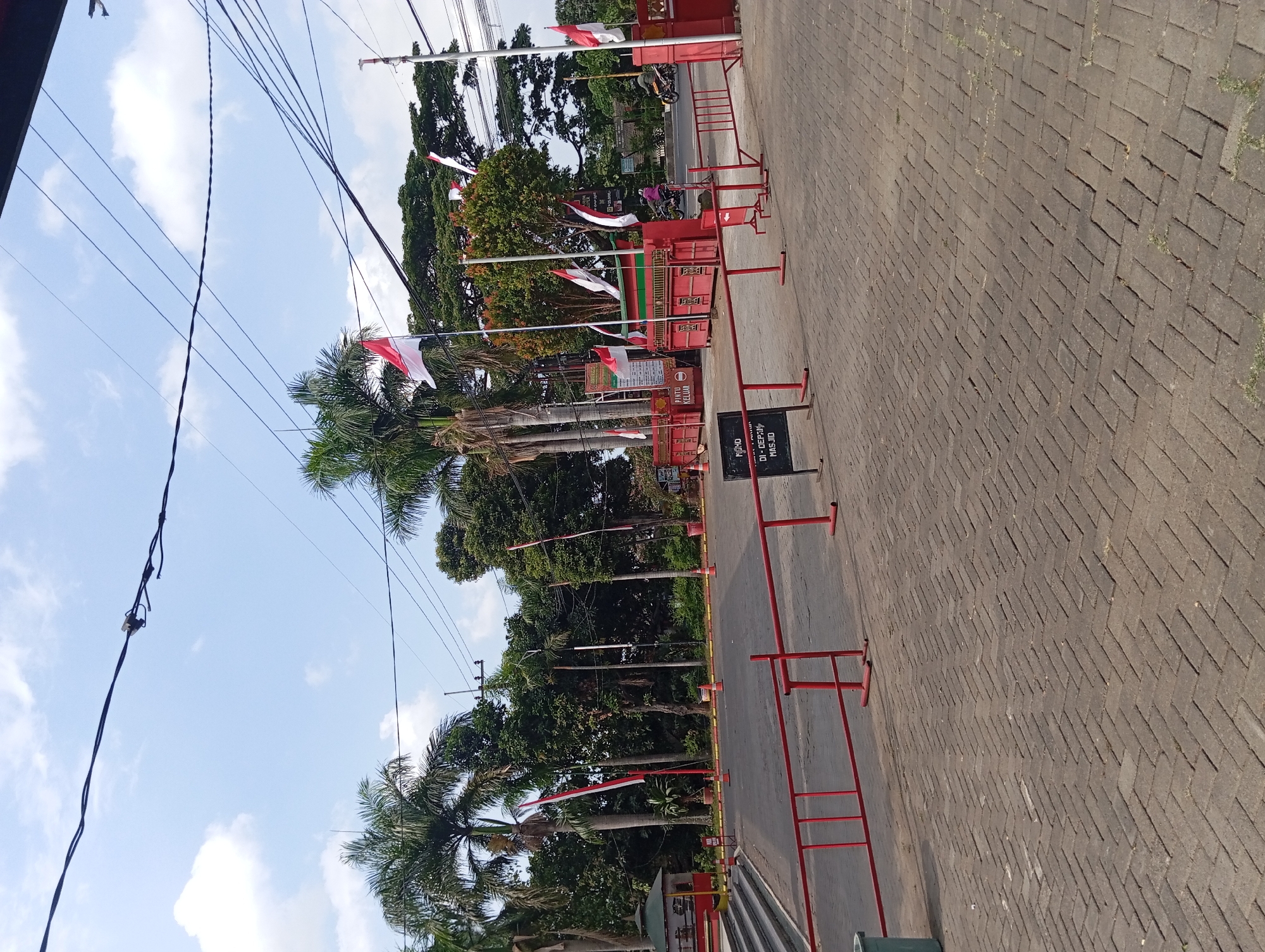 The image shows the entrance to a facility in Pandaan, Indonesia, possibly a museum or park, with a clear blue sky and scattered clouds above. Several tall palm trees and other lush green trees are visible, creating a tropical atmosphere. The entrance is marked by a red gate structure with a sign that reads "Pintu Keluar" (Exit Gate) and "Pintu Masuk" (Entrance Gate). Indonesian flags, red and white, are displayed prominently on poles. A paved walkway with interlocking bricks leads towards the entrance, and red metal barriers are set up on the walkway. A small green trash bin is also visible. In the background, a road is present with a few vehicles, including motorcycles, one of which is carrying two people. The overall scene appears to be a public space on a bright, sunny day.