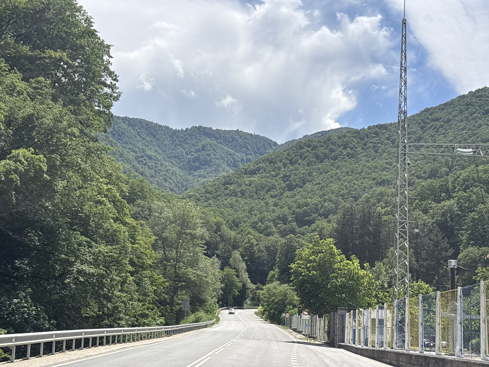 The image shows a road winding through a lush, green mountainous landscape. On the left side of the road, a silver metal guardrail runs alongside, separating the road from a steep, tree-covered slope. The road itself is paved, with a double white line indicating two lanes of traffic. In the distance, a single white car is visible driving away from the camera. The right side of the road features a chain-link fence with colorful panels, and a tall, metallic communication tower stands prominently against the sky. The background is dominated by rolling hills densely covered with trees, leading up to a hazy, sky with scattered clouds. The scene appears to be a scenic route in a rural or mountainous area. This location is the road leading to the Yundola area, near Peshtera, Bulgaria.