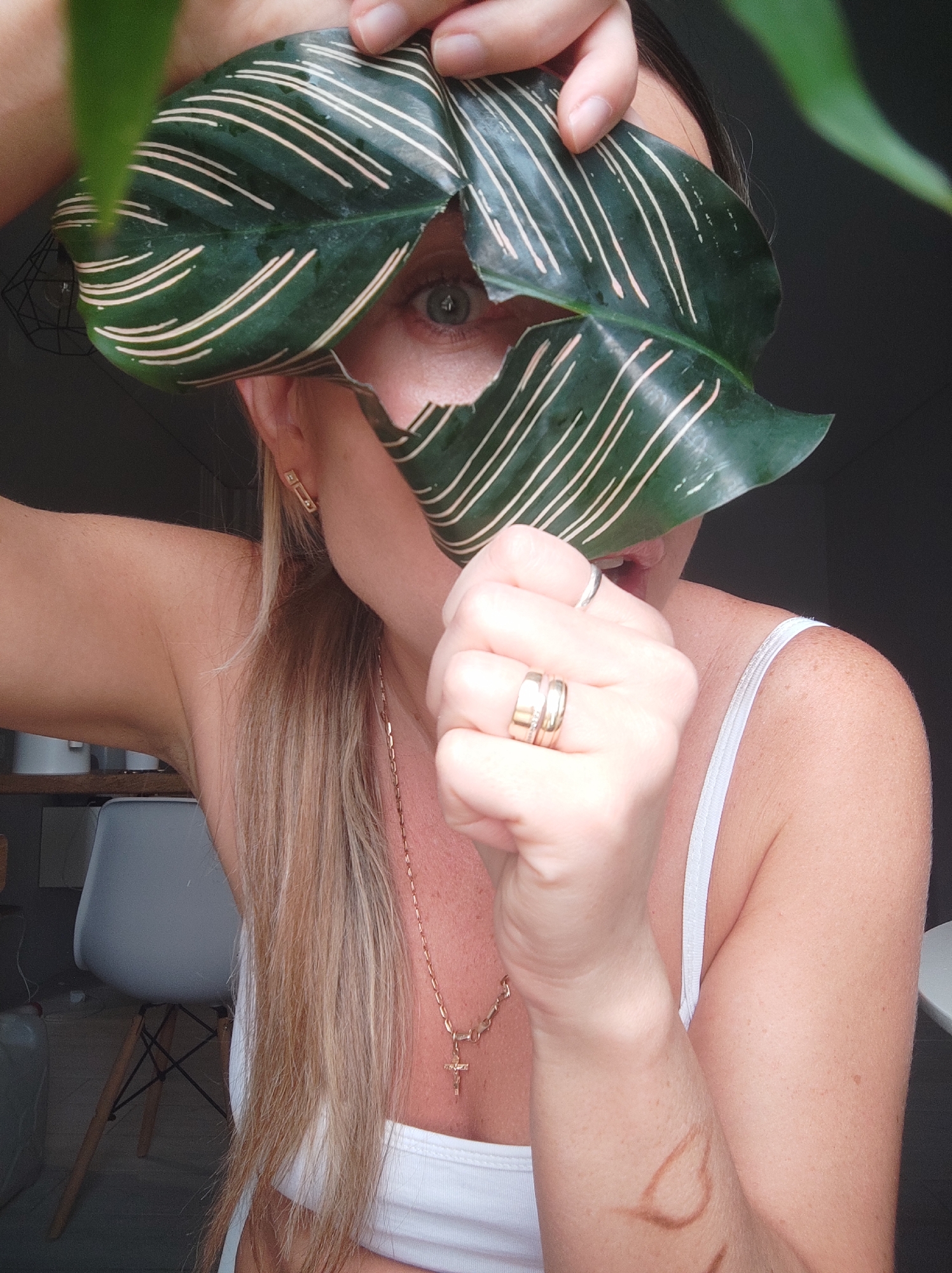 A woman poses indoors, holding a large green leaf with cream-colored stripes in front of her face, partially obscuring it. Only one eye is visible. She is wearing a white tank top and several rings. A gold necklace with a cross pendant is visible around her neck. She has long blonde hair pulled back in a ponytail. A brown heart-shaped mark is on her arm. In the background, there is a white chair and some shelves with white items on them. It is not possible to determine if the location is in Votkinsk, Russia based on the image alone.