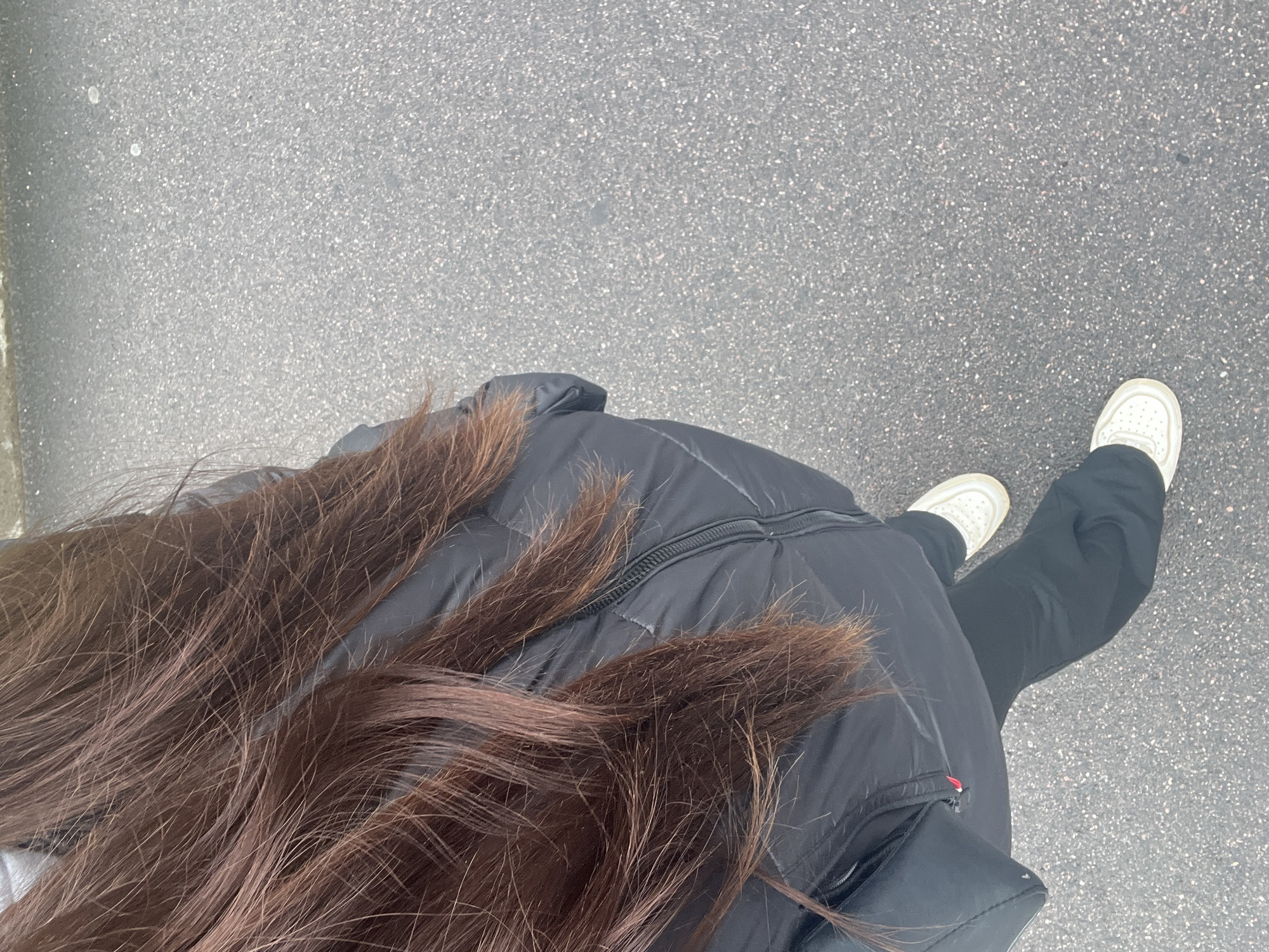 The photograph is taken from an elevated perspective, looking down at a person walking on a gray asphalt surface. The person is wearing a black puffer jacket and dark pants, with white sneakers visible. Long, brown hair is draped over the jacket. The ground is a grainy asphalt. The location and city are not discernable from the image. There are no other people or objects visible. The overall impression is of a casual, everyday scene of a person walking.