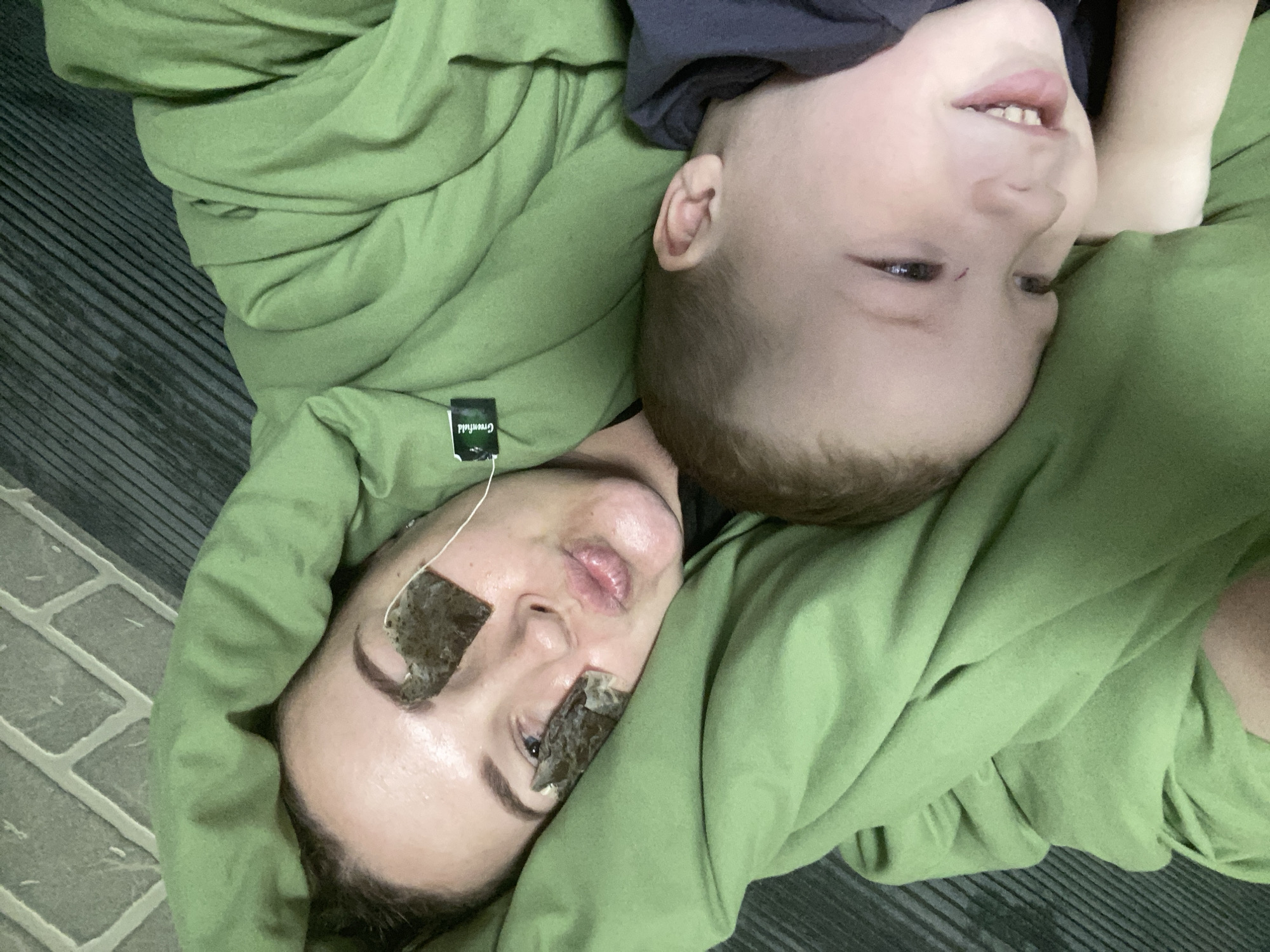 A woman and a young boy are lying on what appears to be a floor covered in a green blanket. The woman has tea bags on her eyes. The image was likely taken indoors, but there is not enough information to confirm if the location is Sochi, Russia.