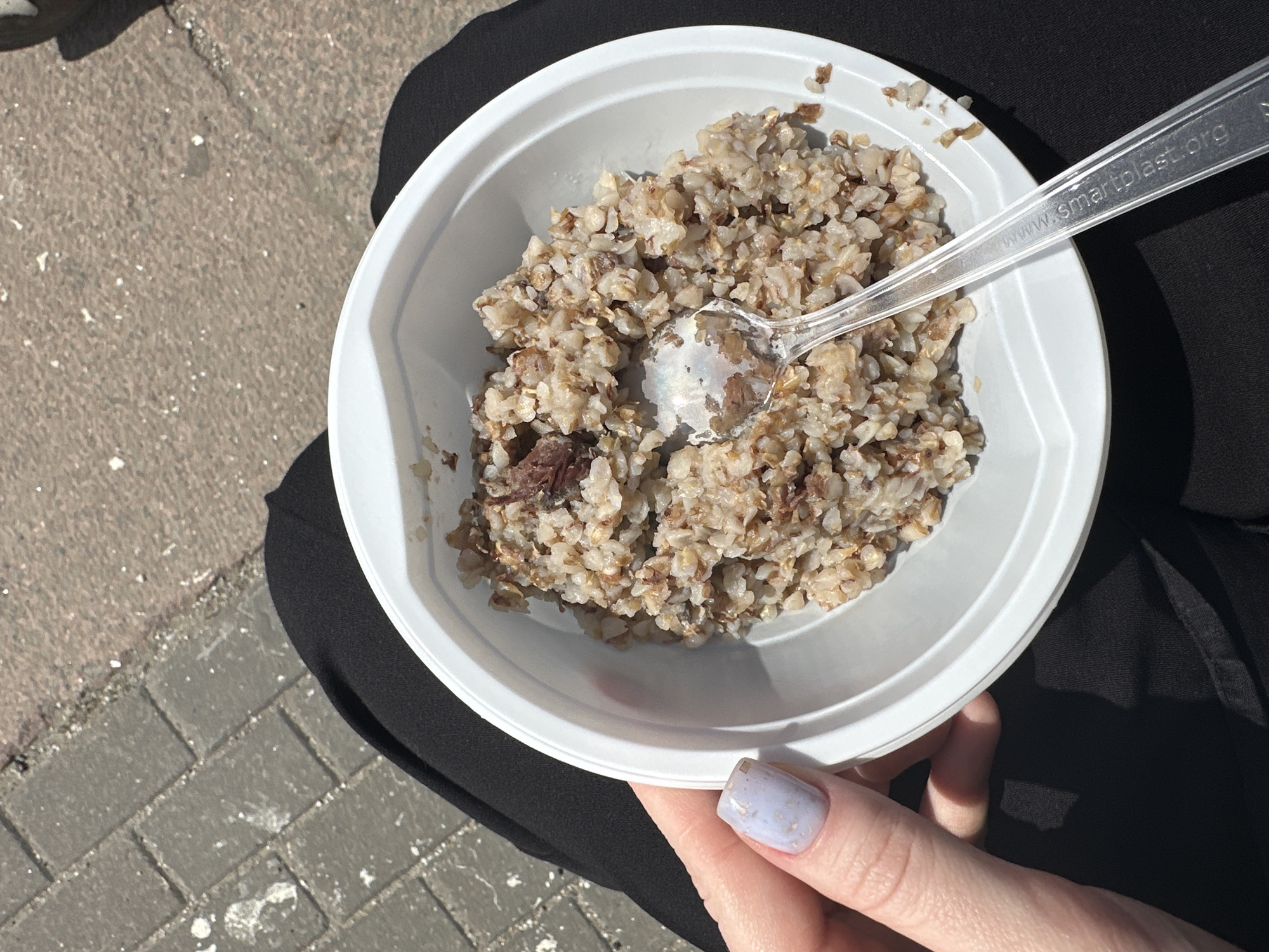 An overhead shot depicts a white plastic bowl containing buckwheat cereal with pieces of meat, held by a person, presumably sitting outdoors in Moscow, Russia. A transparent plastic spoon rests inside the bowl, its handle printed with "www.smartplast.org." The person's hand, visible in the lower right, features a fingernail painted with a light blue polish. The person is wearing black pants. Patches of sunlight and shadow are visible on the bowl, the pants, and the gray brick sidewalk in the background.