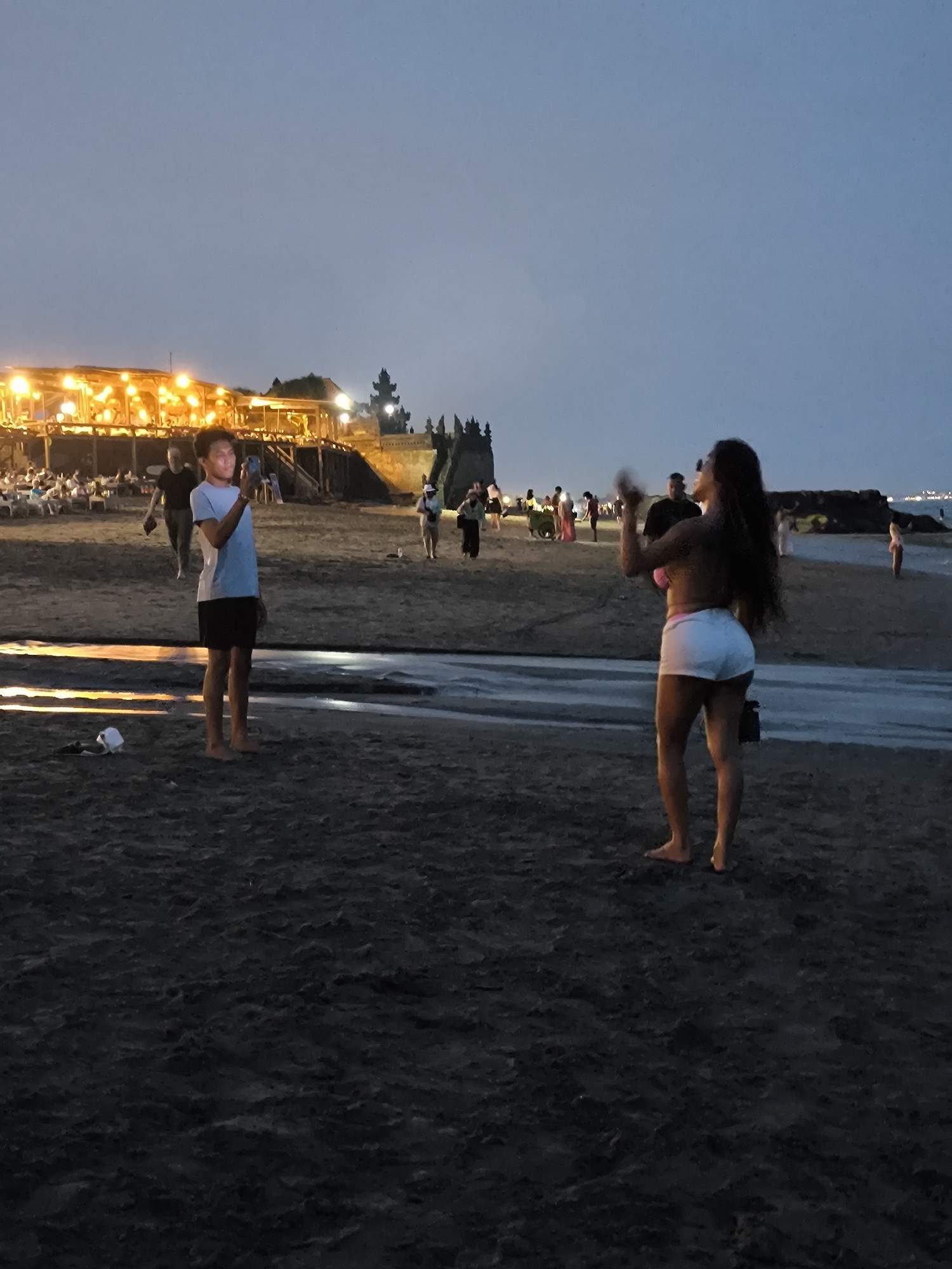 The image shows a beach scene at dusk in Canggu, Indonesia. In the foreground, a young man stands barefoot, holding a smartphone as if taking a picture. He is wearing a light blue t-shirt and dark shorts. To his right and slightly behind him, a woman in a pink bikini top and white shorts stands with her back to the camera, holding her arms up. A small black bag is beside her feet.
In the background, several other people are visible along the beach. A lighted building stands on a raised area, suggesting a restaurant or bar. Water flows between the foreground and the background, indicating a small stream or tidal area. The sky is overcast.