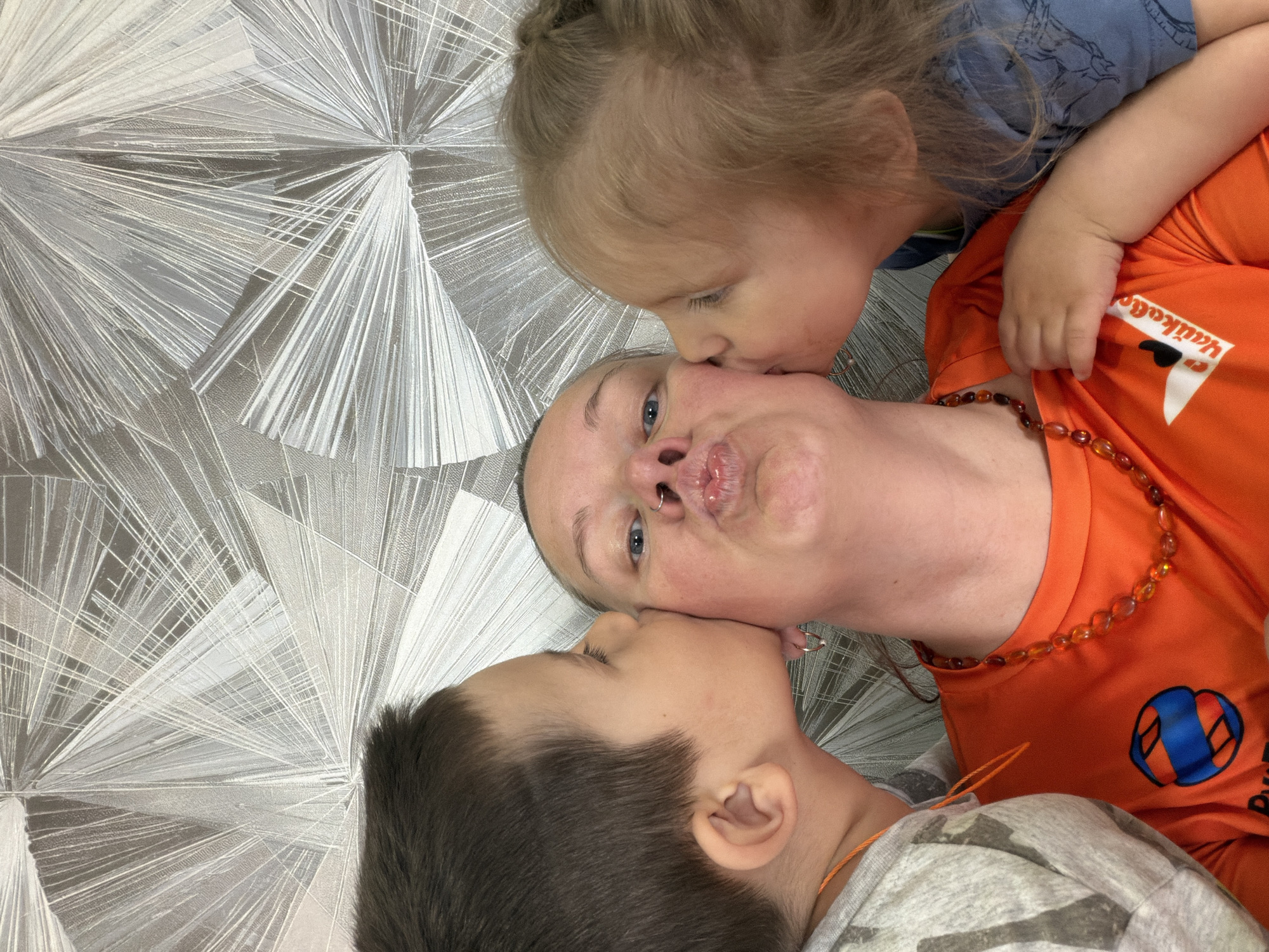 The image shows an adult female lying on a surface with a patterned design. She is wearing an orange t-shirt with an orange beaded necklace. Two children are positioned next to her, appearing to kiss her face. The child on the left has dark hair and wears a grey patterned shirt. The child on the right has blonde hair and wears a blue shirt. The location is Chaikovsky, Russia.