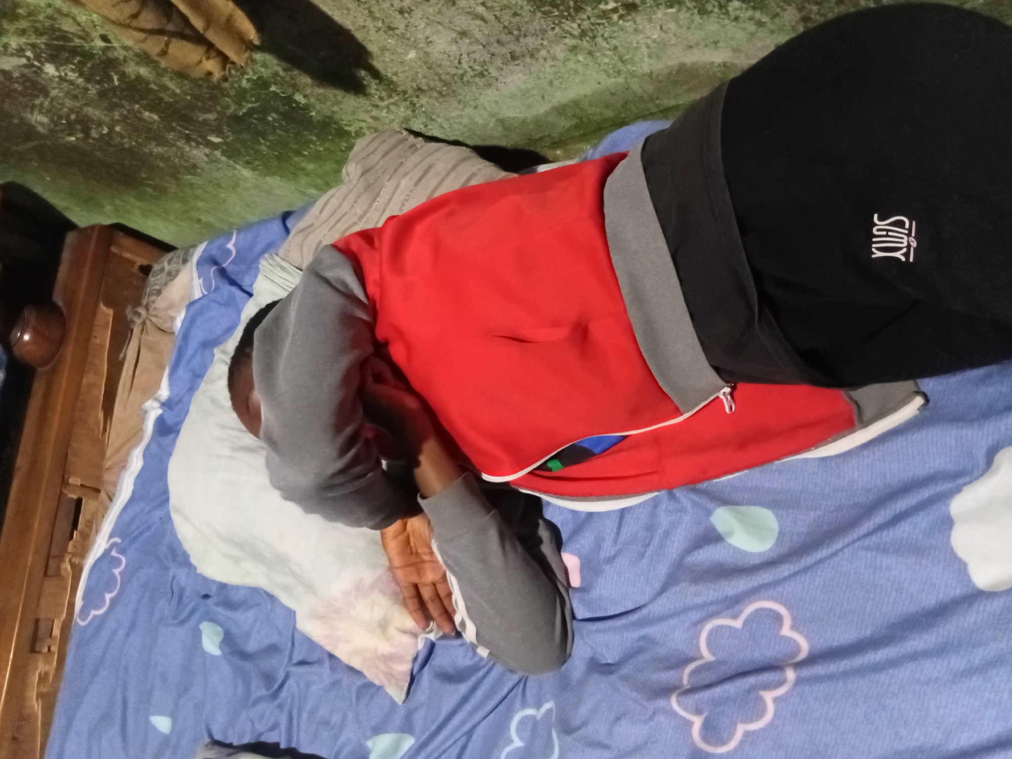 A person is sleeping on a bed covered with a blue duvet featuring cloud and raindrop patterns. The person is wearing a red and gray zip-up jacket and black pants with a logo on the left thigh. Their head is resting on a pillow, and their arms are wrapped around their head. The bed is situated in a room with a wooden headboard and a textured green floor. The scene appears to be in Nkpor, Nigeria.