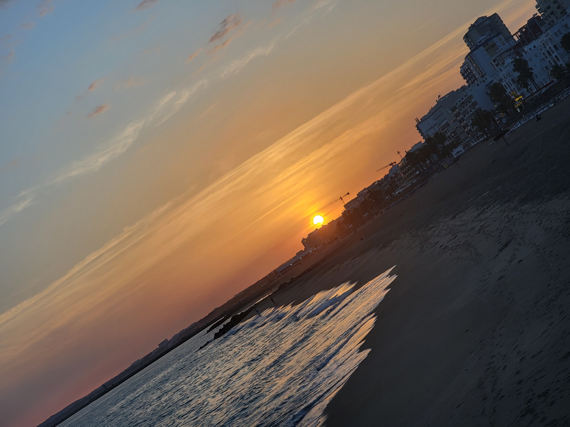 The image depicts a wide-angle view of a beach and cityscape at sunset in Quarteira, Portugal. The sun, a bright orange orb, is low on the horizon, casting a warm glow across the sky and reflecting on the ocean. The sky transitions from a pale blue at the top to hues of orange and pink near the horizon, with wispy clouds scattered throughout.
The foreground features a dark sandy beach with gentle waves lapping at the shore, creating white foam. The water appears dark due to the low light conditions. In the midground, a line of buildings forms the Quarteira cityscape, characterized by multi-story residential or hotel structures with a modern architectural style, predominantly white or light-colored. Several palm trees are visible amongst the buildings, suggesting a coastal resort town. Construction cranes are also present, indicating ongoing development in the area.
The image is taken from a slightly elevated perspective, looking out towards the sea and the setting sun. The overall impression is one of a tranquil evening scene at a popular tourist destination. There are a few indistinct figures on the beach, suggesting a low level of activity. The tilt of the image suggests it was captured with the camera held vertically and then rotated, or the scene itself is on an incline.