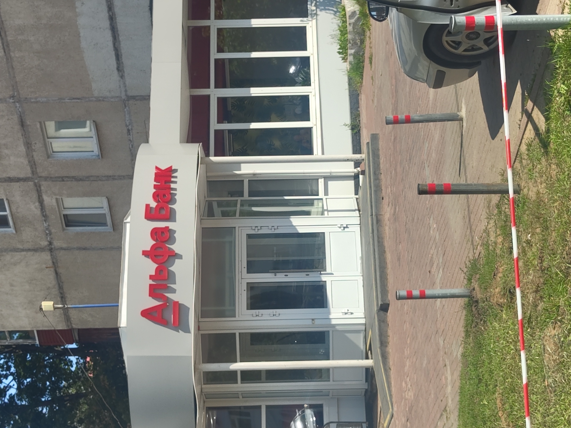 In Ramenskoye, Russia, an Alfa-Bank branch is visible, identified by red lettering on the building's facade. The bank features a modern glass entrance with double doors, flanked by large windows. The surrounding area is paved with brickwork and includes patches of green grass. Several bollards, some with red and white reflective stripes, are placed along the sidewalk. A light-colored car is parked in front of the bank, its driver's door open. The bank is situated in front of a concrete apartment building, suggesting an urban or suburban setting. The sky is clear and bright, indicating daytime.
