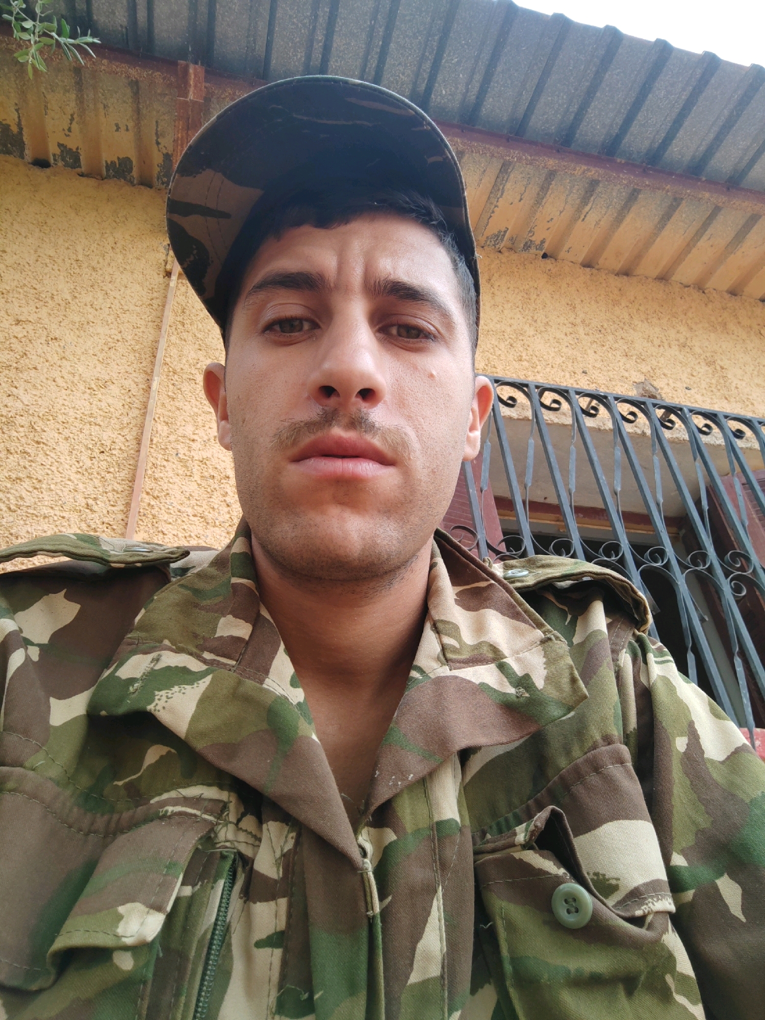 In Mougheul, Algeria, a young man with a mustache is captured in a close-up, waist-up selfie. He wears a camouflage military uniform and a matching cap, positioned forward on his head. His gaze is directed towards the camera. Behind him, a textured yellow wall is visible, with a portion of a dark metal fence or railing to his right, suggesting an outdoor or semi-outdoor setting. The background also includes a corrugated metal roof and some greenery peeking from the top left. The lighting appears to be natural daylight.